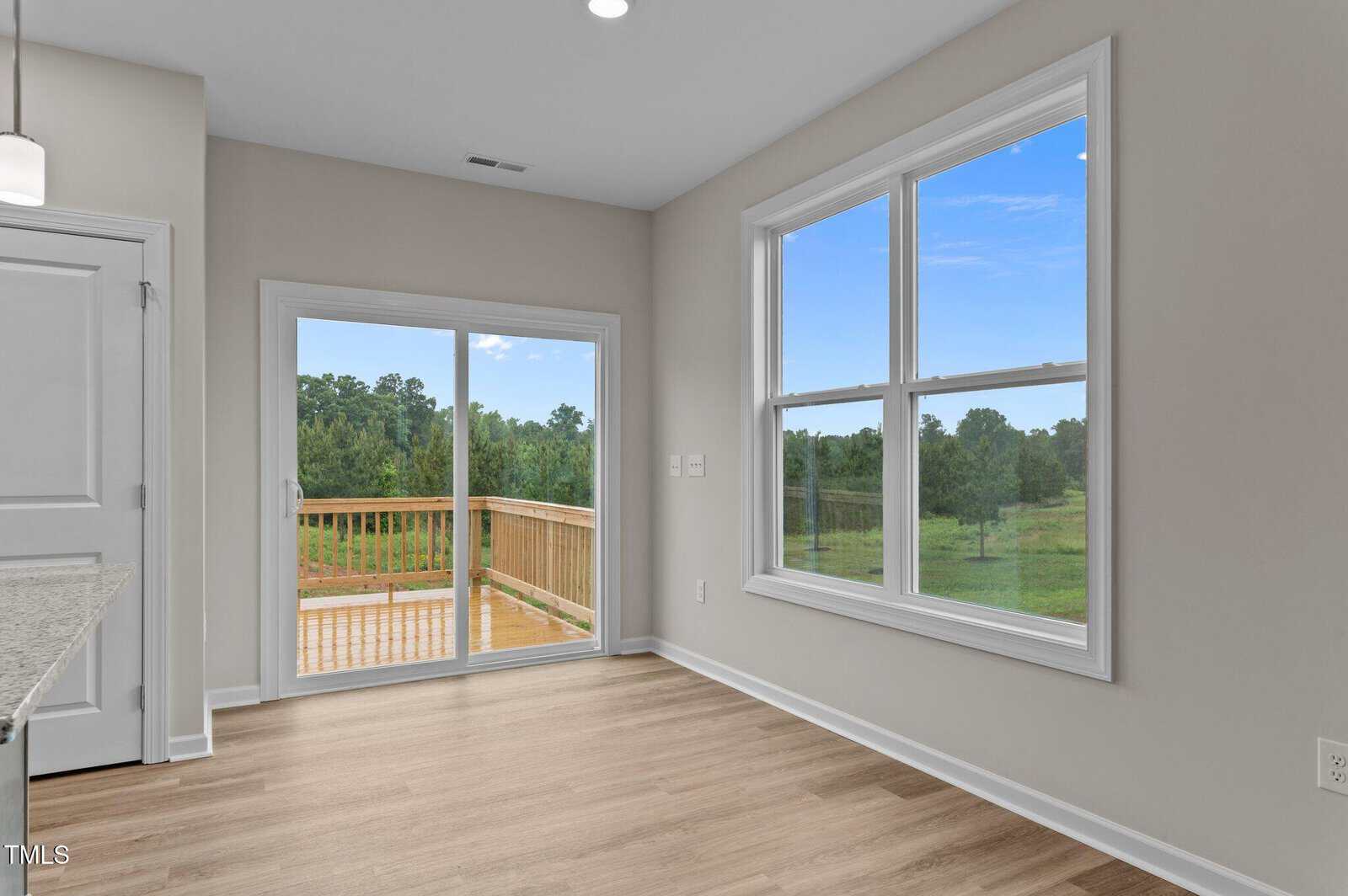 A bright room with light wood floors, beige walls, a large window, and sliding glass doors leading to a wooden deck. Trees and greenery are visible outside under a blue sky.