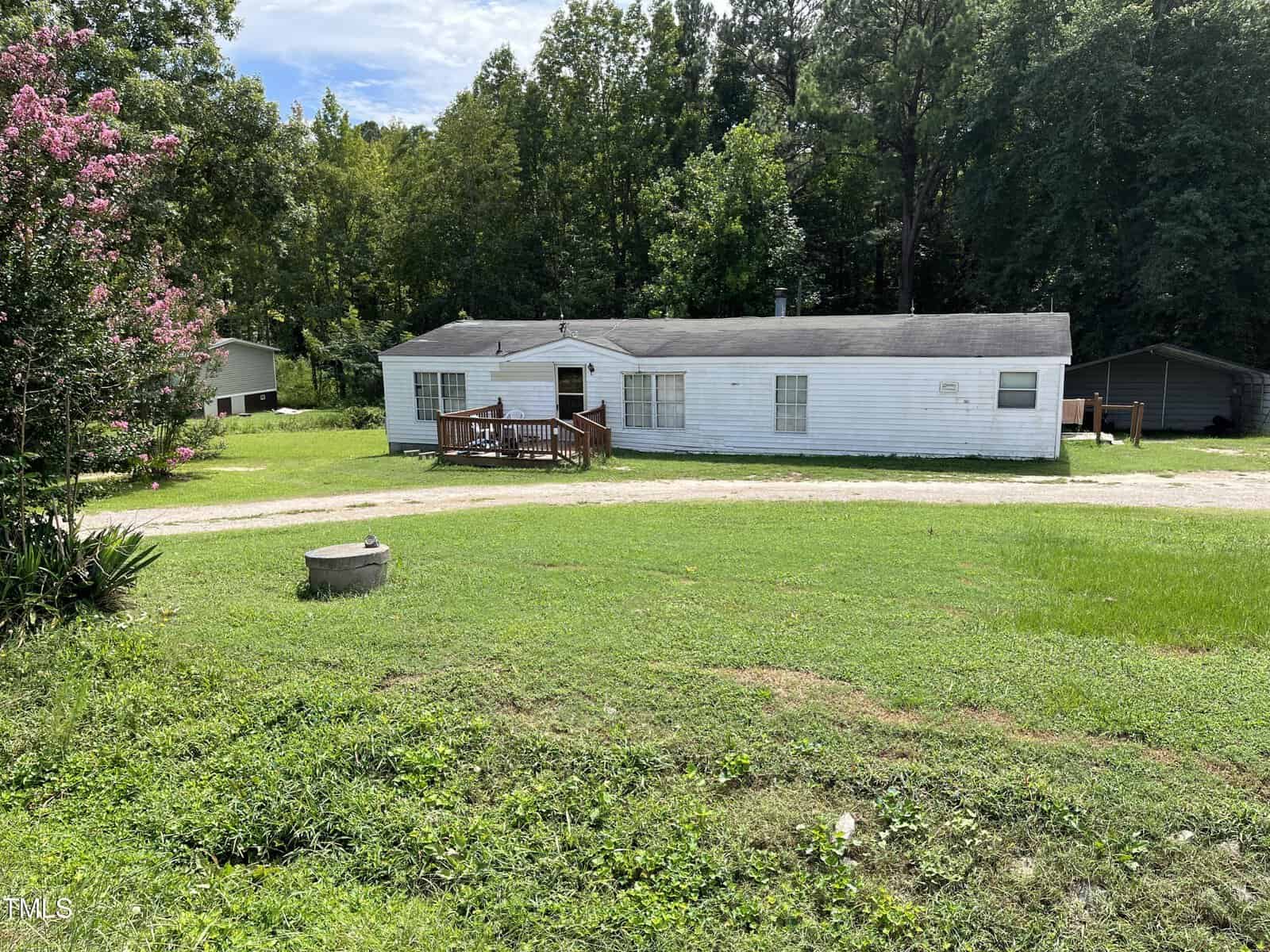 A single-story white house with a small wooden deck sits on a large grassy lawn at 76 Sutton Road, Louisburg. There is a gravel driveway and fire pit in front, with trees and another building in the background. Sky is partly cloudy.