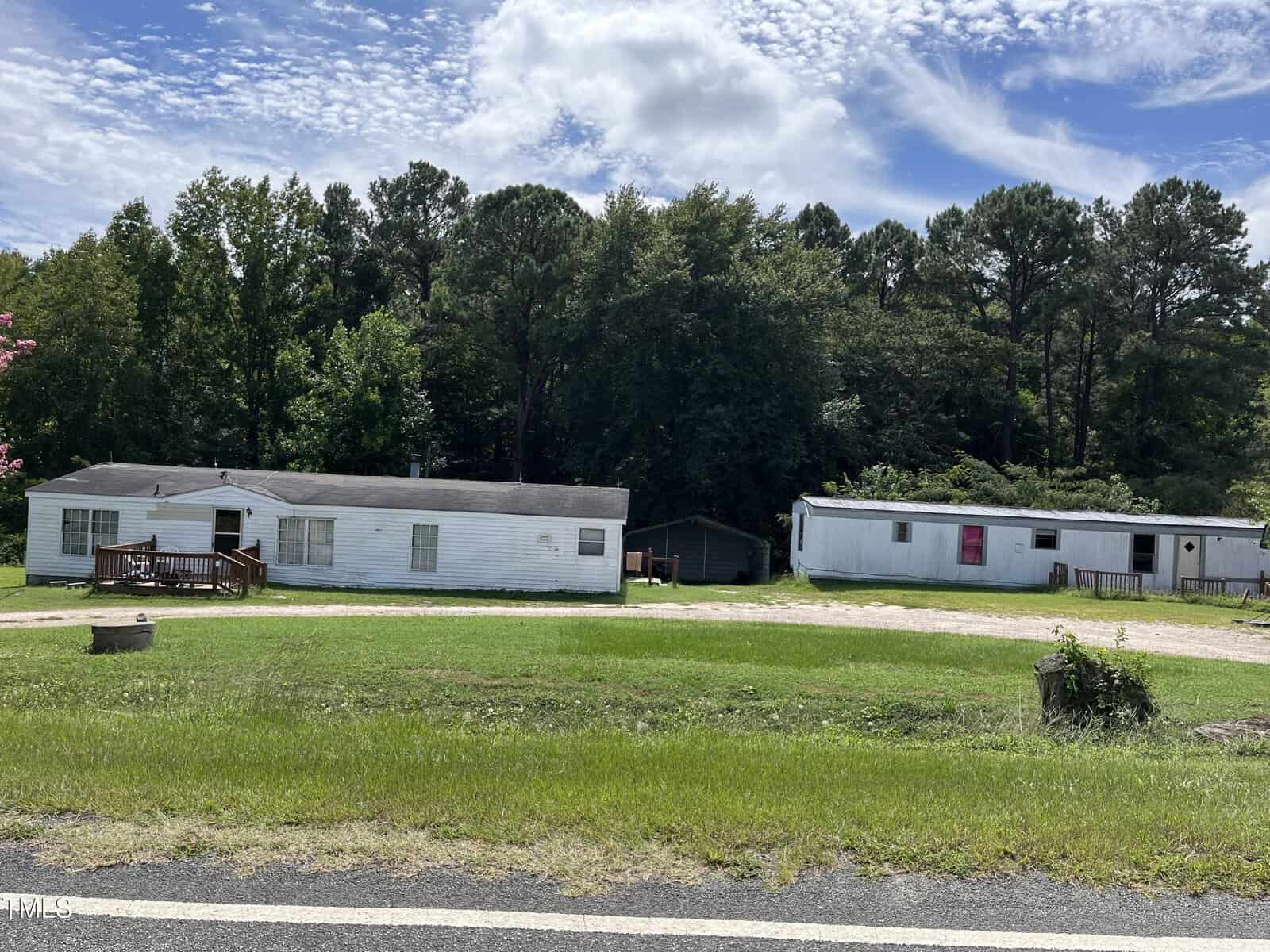 Two white mobile homes with weathered exteriors sit on a grassy lot near a gravel driveway, backed by dense green trees under a partly cloudy sky. A road runs across the foreground.