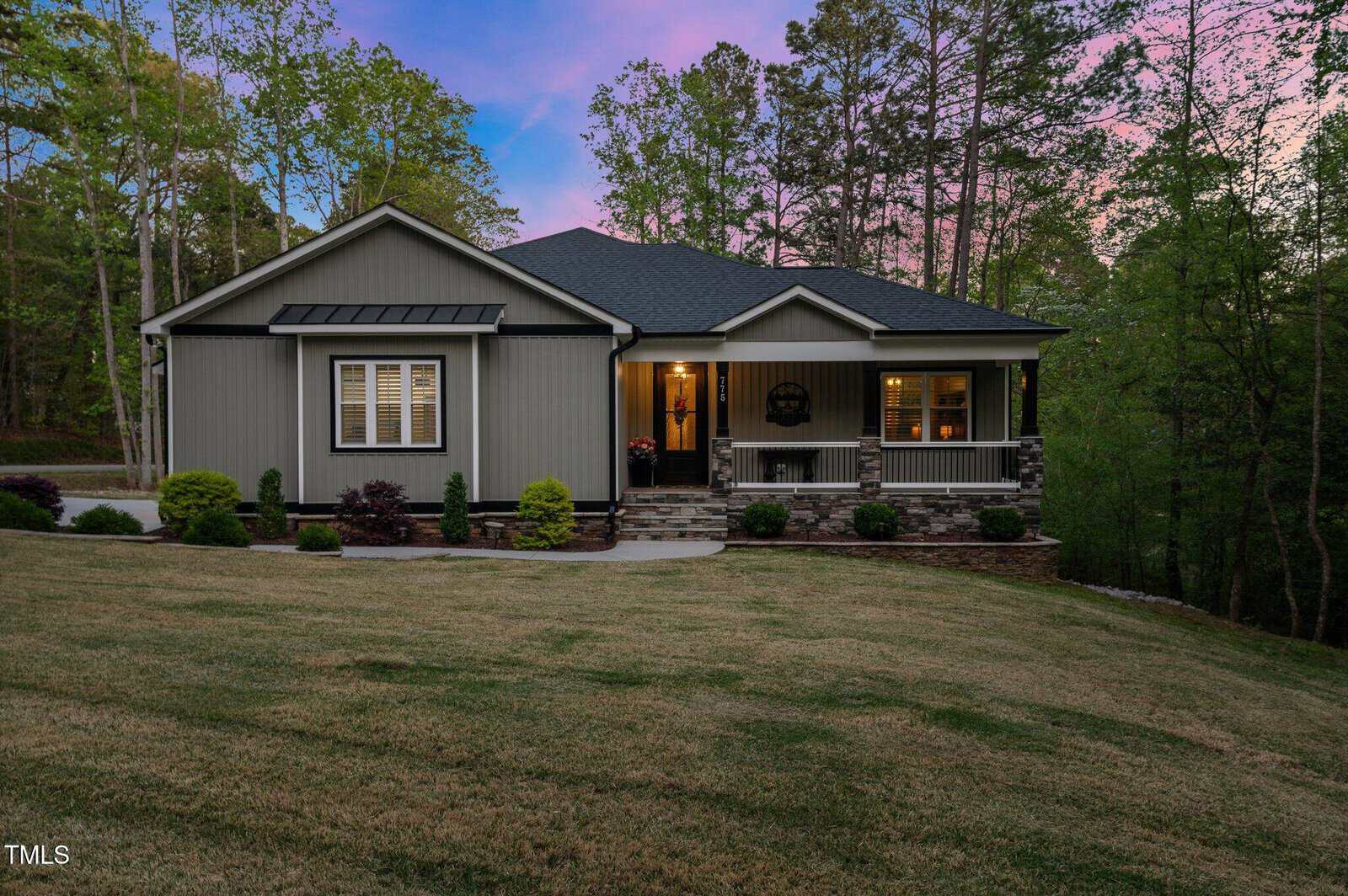 Single-story house with gray siding, stone accents, and a covered front porch. The lawn is sloped and manicured, surrounded by trees. The house is lit from inside, and the sky is tinged with pink and blue at dusk.