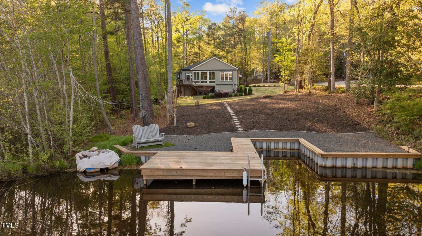 A small house sits among trees at the end of a gravel path, leading from a wooden dock on a calm lake at 775 Sagamore Drive in Louisburg. Two Adirondack chairs face the water, and a canoe is tied near the dock. The area is surrounded by woods.
