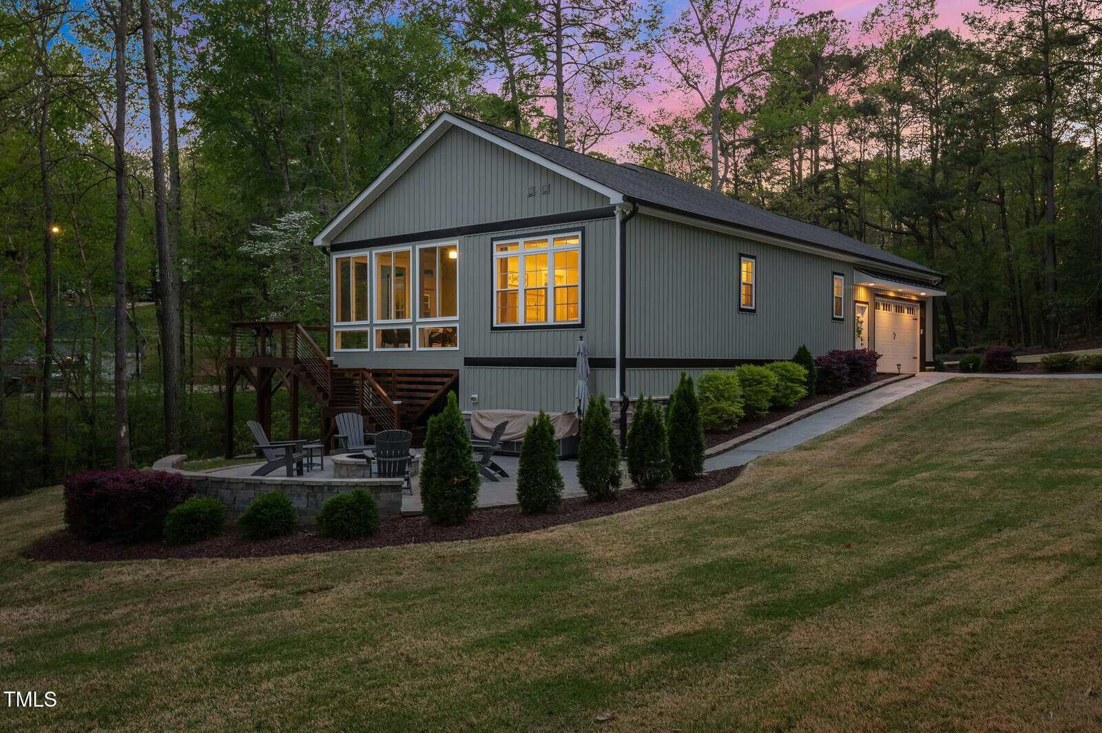 Single-story house with large windows lit from within at dusk, surrounded by trees. A landscaped lawn has a paved patio area with chairs and a fire pit. The driveway leads to an attached garage on the right side.