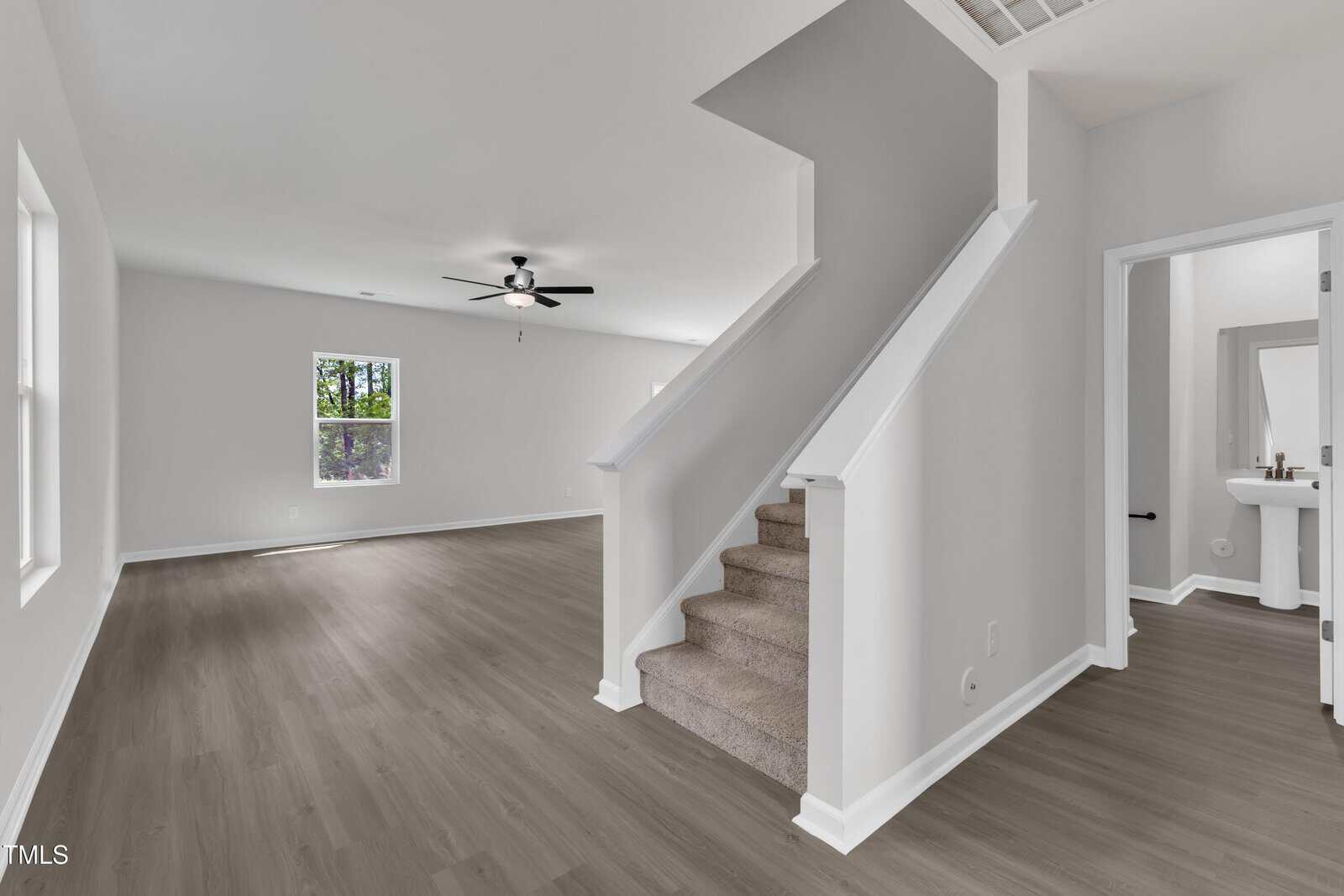 A modern, empty living room with light gray walls, wood-look flooring, a ceiling fan, and a carpeted staircase leading up. A small bathroom with a pedestal sink and mirror is visible to the right.