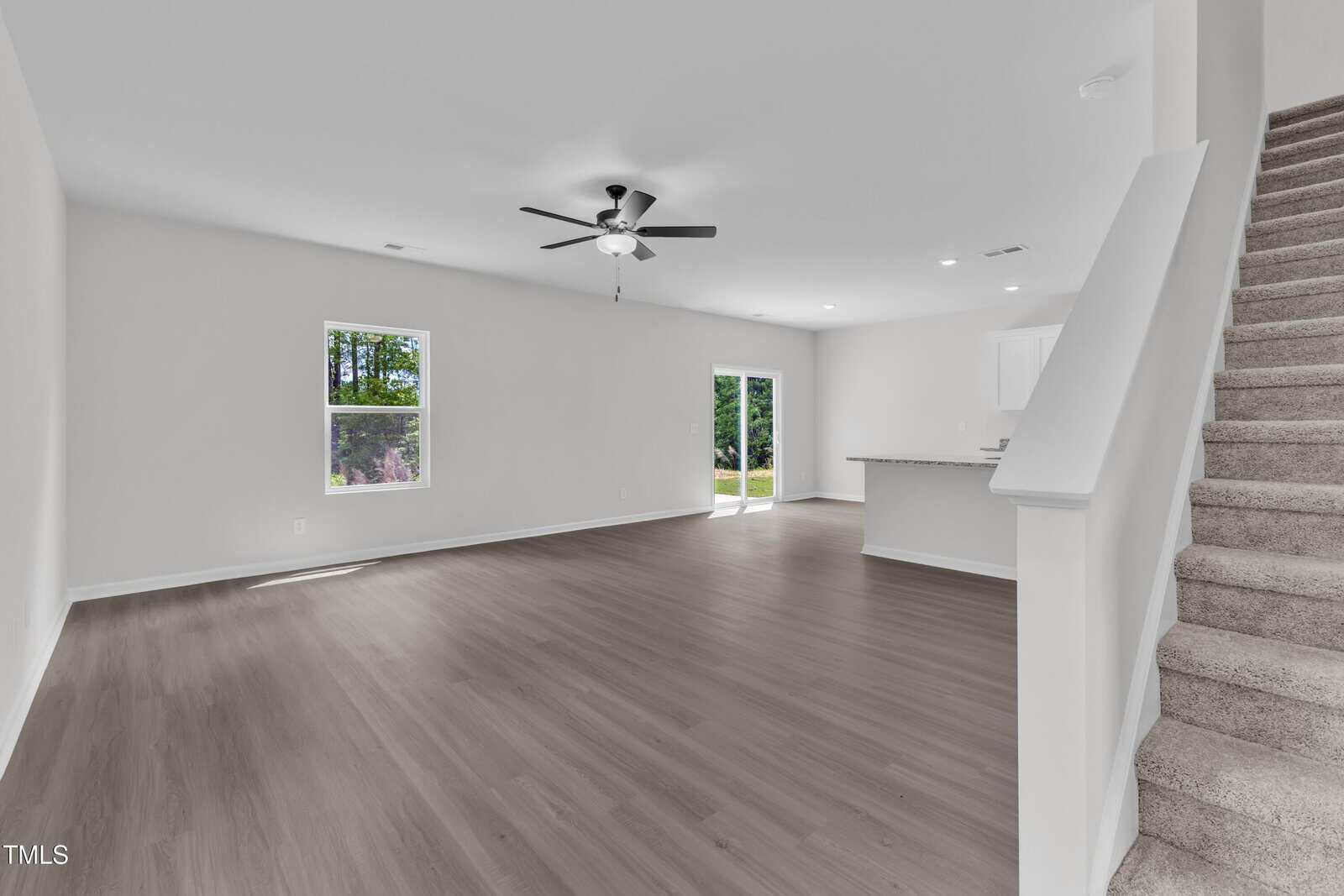 Open-concept living area with light-colored walls and wood flooring, featuring a ceiling fan, two windows, sliding glass doors, a kitchen with a breakfast bar, and carpeted stairs leading to the upper floor.
