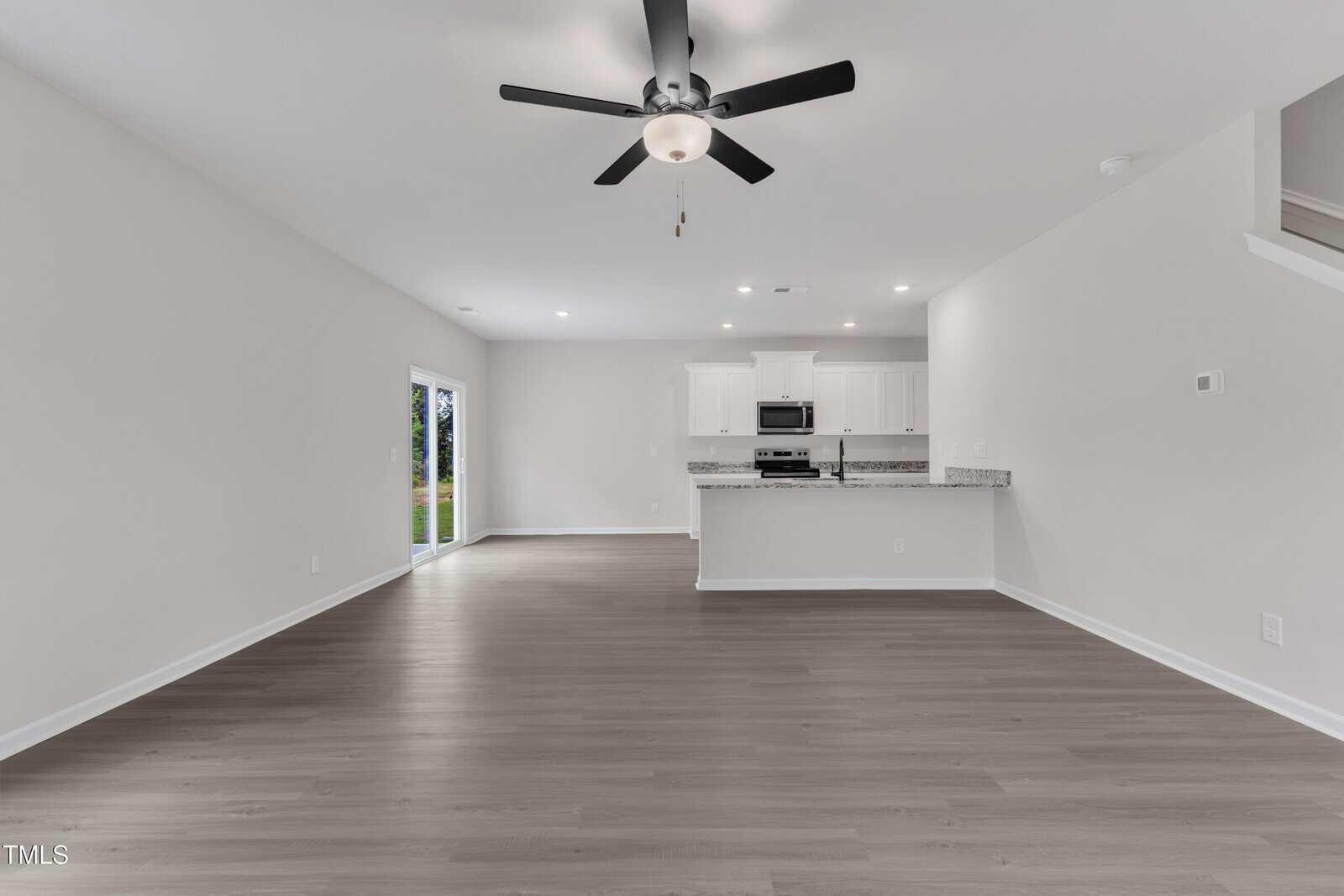 Unfurnished open-plan living area with light-colored walls and wood-look flooring, a ceiling fan, recessed lighting, and a kitchen with white cabinets and granite countertops. Sliding doors lead to the outside.
