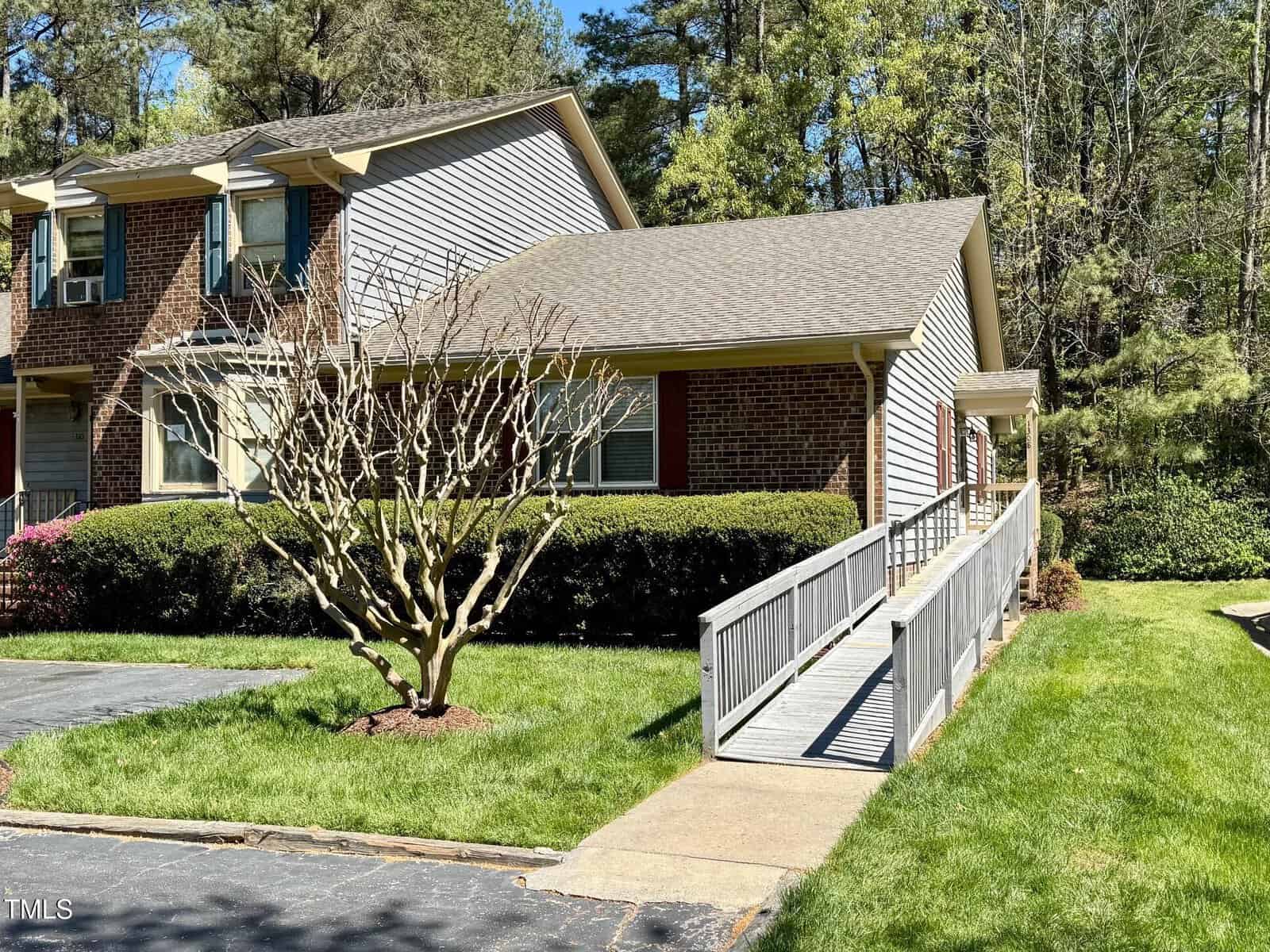 At 1308 Oakridge Avenue Unit 3, this charming brick townhouse in Henderson boasts a beige and white exterior with a wooden ramp leading to the inviting entrance. A manicured hedge and a small, leafless tree lie beside the lush green grass surrounding it.