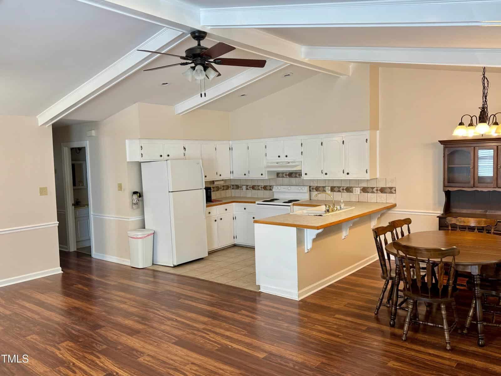 A kitchen with white cabinets, a refrigerator, stove, and dishwasher. There's a wooden dining table with six chairs. The area has hardwood flooring and a ceiling fan. The walls are painted in beige, and there's a light fixture above the table.