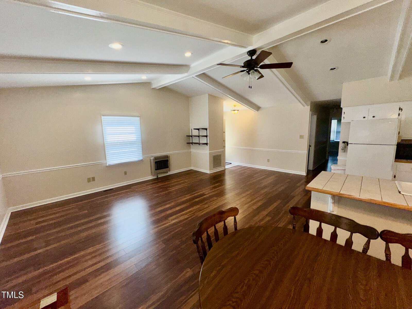 A spacious living area with wood flooring and white walls. A ceiling fan is mounted above. A window with blinds is on one wall. The open kitchen area features a countertop and a white refrigerator. A small dining table with chairs is in the foreground.