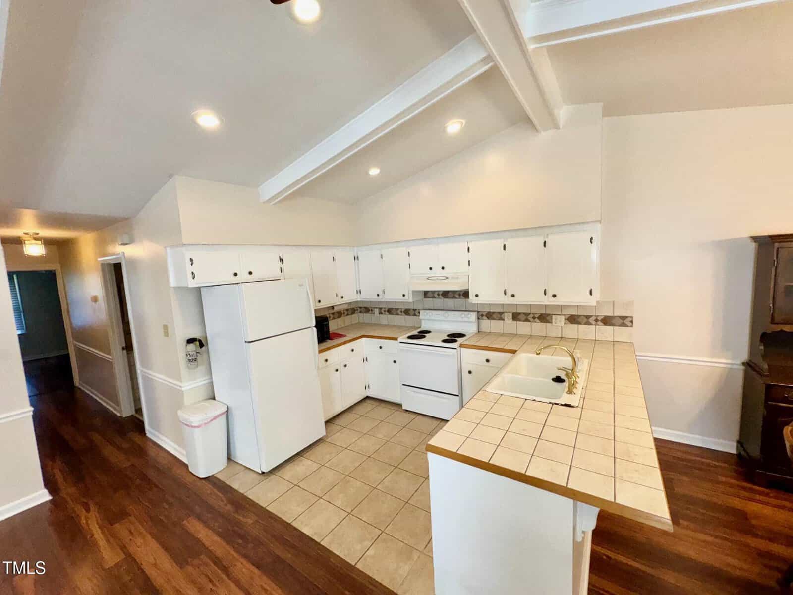 A kitchen with white cabinets and appliances, including a stove, oven, fridge, and microwave. The countertop is tiled, and the floor combines wood and tile. Recessed lighting is installed in the ceiling, and a hallway is visible on the left.