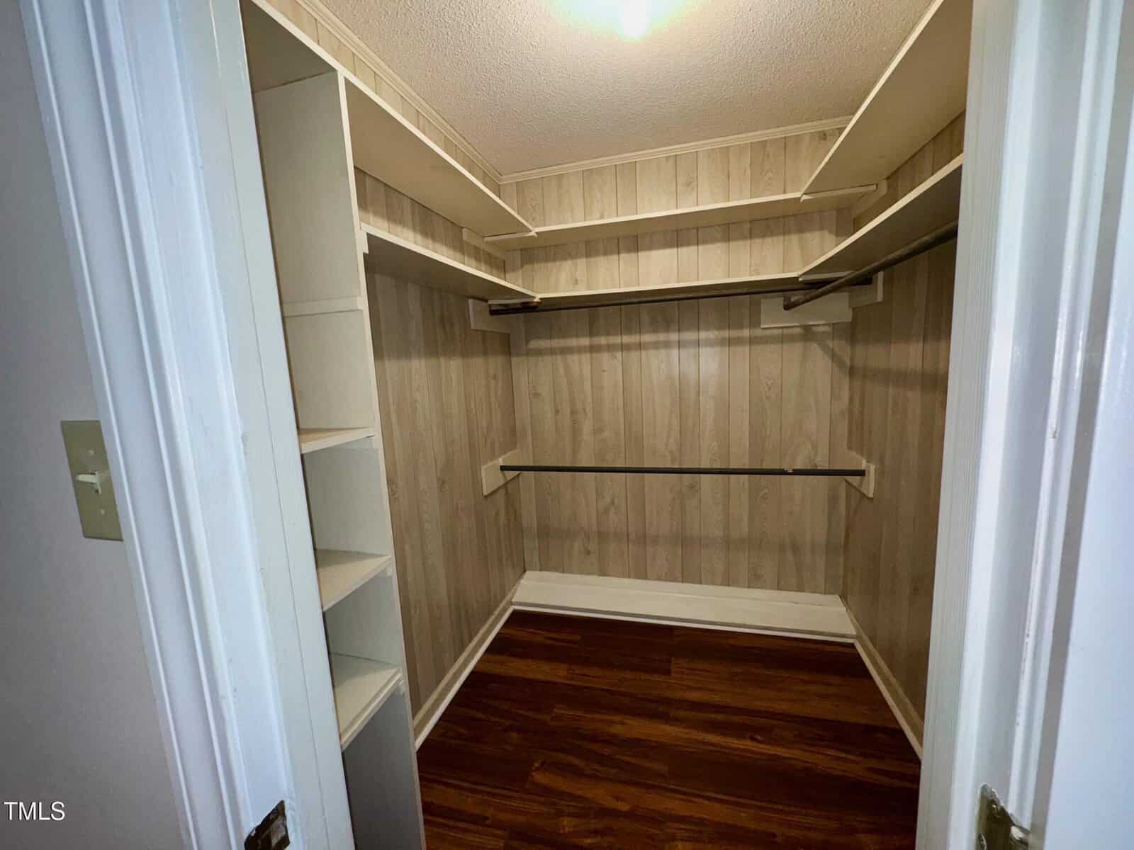 Walk-in closet with wood-patterned paneling and hardwood floor. Features multiple shelves and hanging rods on three walls, providing ample storage space. Ceiling light fixture illuminates the area.