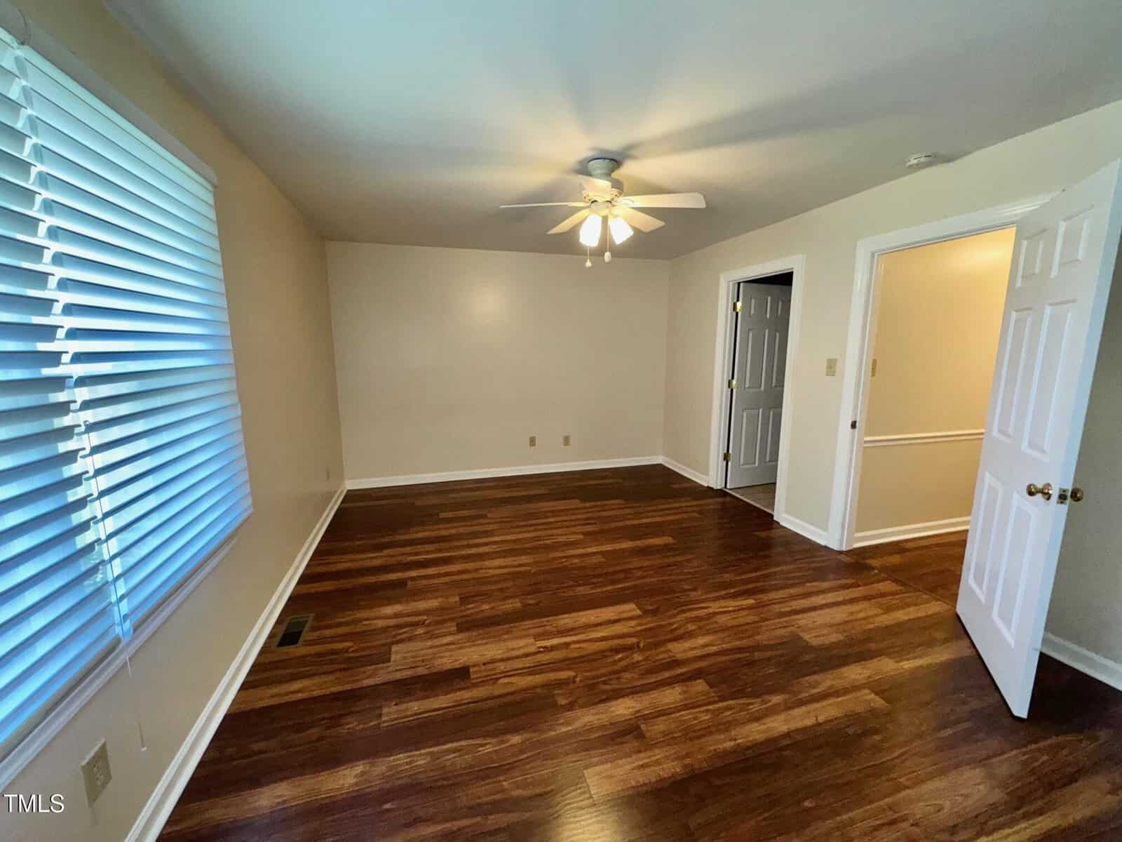 An empty room with wooden floors, beige walls, and a ceiling fan with lights. Two doorways, one with an open white door, are visible. A large window with closed blinds is on the left wall.