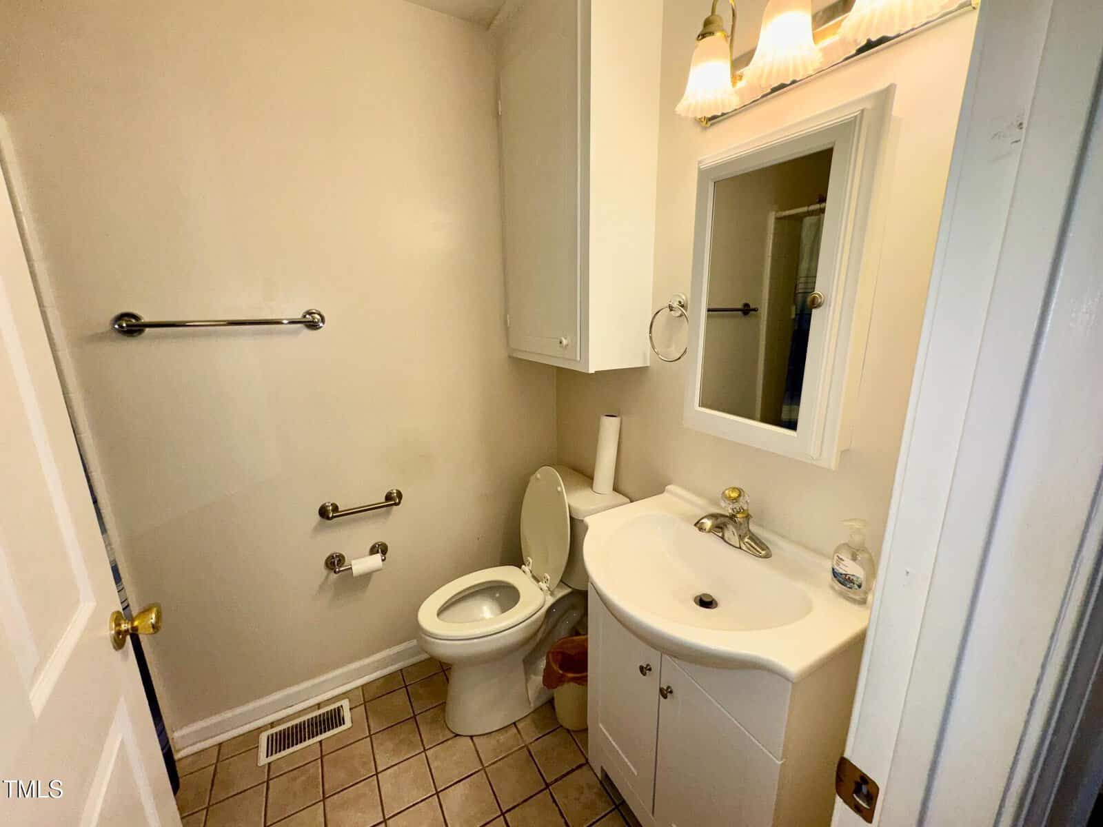 Small bathroom with beige walls and tiled floor. Features a toilet, white vanity with sink, mirror above, light fixture, and towel bars. Soap dispenser and toilet paper are visible. A ceiling vent is also present.