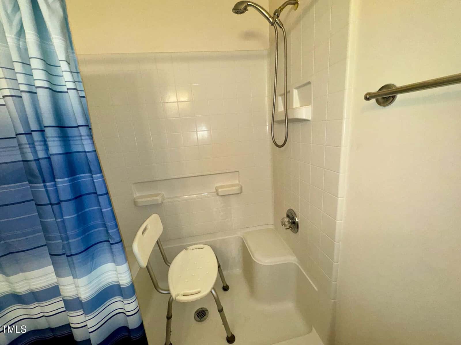 A bathroom with a tiled shower area featuring a blue and white striped curtain. Inside is a white shower chair with a backrest. The wall has built-in shelves, and a silver faucet and showerhead are installed. A towel bar is mounted outside the shower.