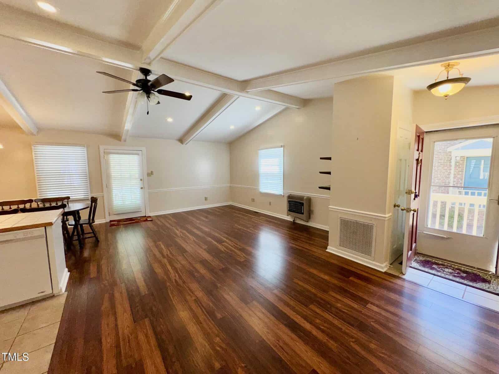 A spacious living area with wooden flooring and white walls. The room features exposed beams, a ceiling fan, and recessed lighting. Windows and an open door provide natural light. A small table with chairs is visible in the corner.