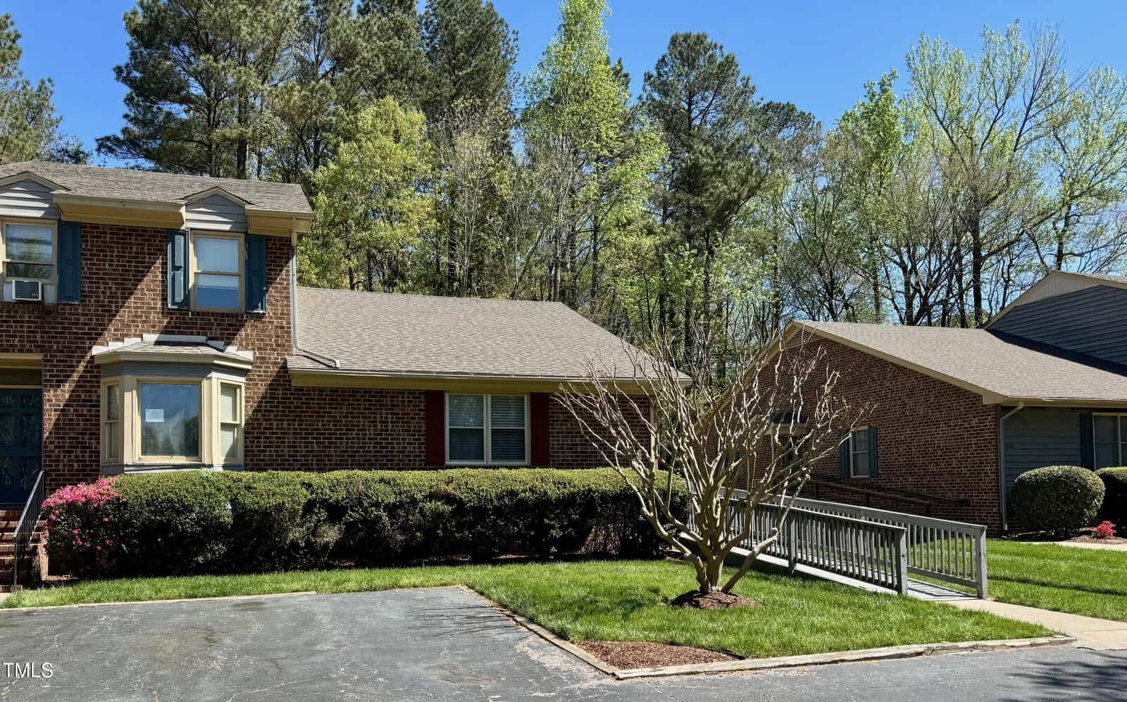 A brick duplex in a suburban setting with a sloped shingle roof and a wheelchair ramp leading to the front door. Bushes and a tree are in the foreground, while trees with new leaves are in the background under a clear blue sky.