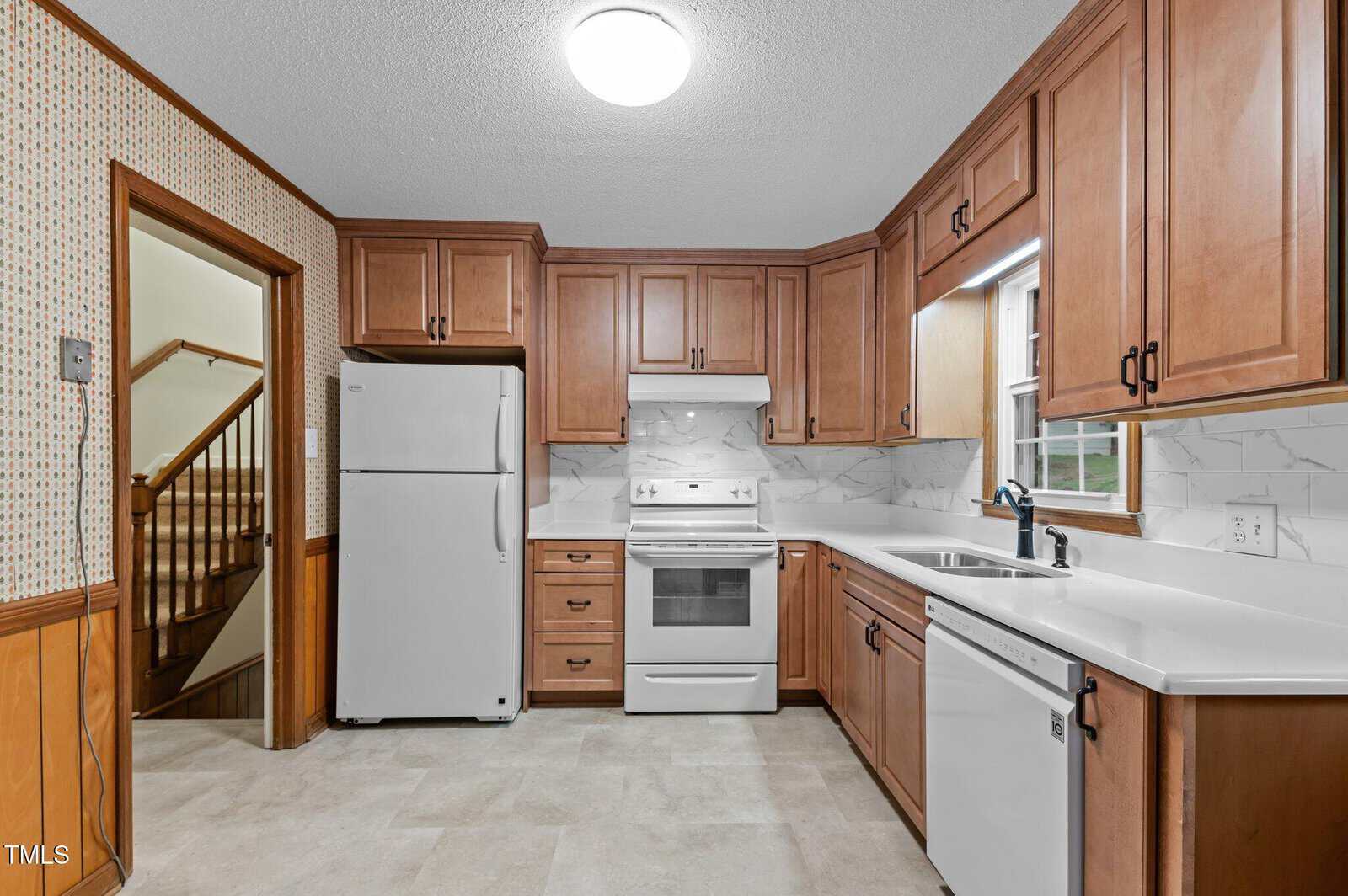 A kitchen with wooden cabinets, a white refrigerator, an electric stove, and a dishwasher. The countertops are white, and the walls have a light pattern. A window above the sink allows natural light in, and a staircase is visible through the doorway.