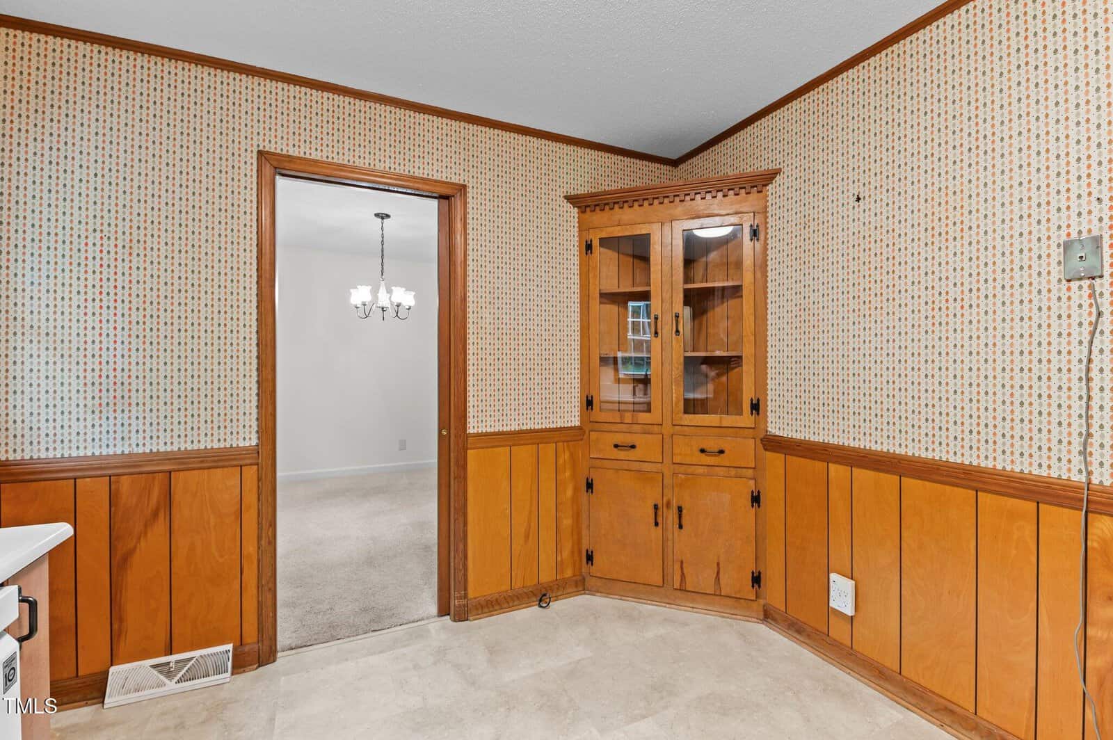 A kitchen corner features wood paneling and a built-in wooden cabinet with glass doors displaying small items. The beige walls have a patterned design. A doorway on the left leads to a carpeted room with a chandelier. The floor is tiled.