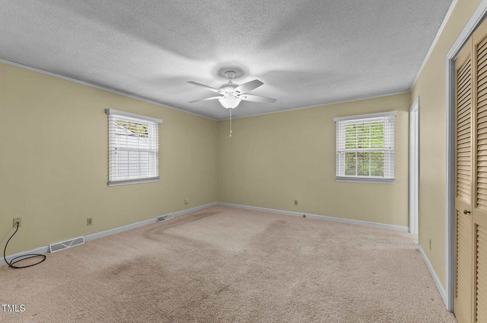 A spacious, empty bedroom with beige carpet, light yellow walls, two windows with white blinds, a ceiling fan with lights, and double closet doors. An electrical cord lies on the floor near the wall.