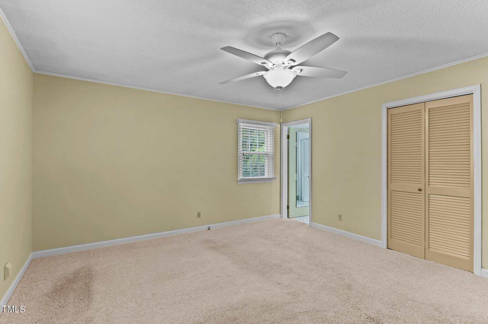 A spacious, empty bedroom with beige carpet, light yellow walls, a white ceiling fan, a window with blinds, a closet with louvered doors, and a doorway leading to a bathroom.