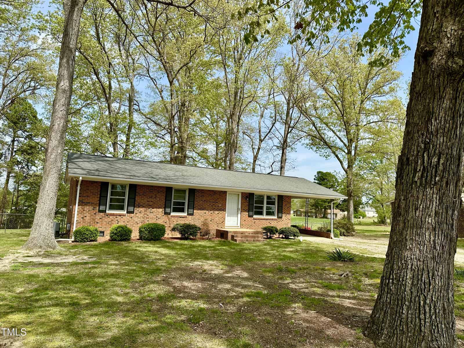 A small, single-story brick house on 1031 Jackson Royster Road in Henderson features a gray roof amid tall trees on a spacious green lawn. The house has a simple white door, several windows, and a short set of steps leading to the entrance. The sky is clear and sunny.
