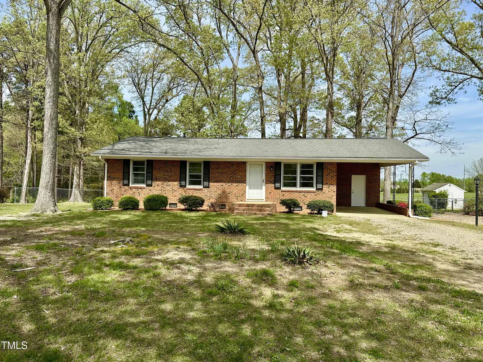 Single-story brick house with a gray shingled roof, surrounded by trees. The house has a white door, several front windows, and a small porch area. The yard is mostly grassy with patches of dirt and some low shrubs near the house.