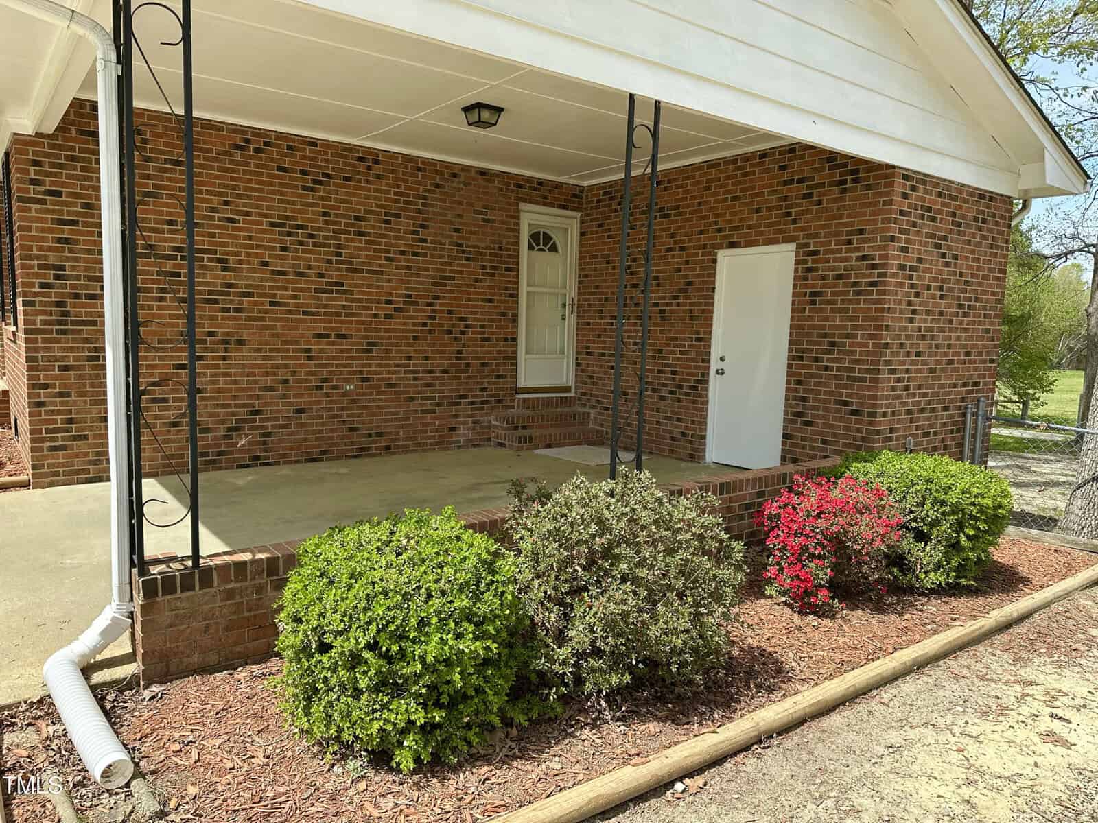 A brick house with a covered entryway and black metal supports. The porch is surrounded by a low brick wall. In front, there are trimmed green and red shrubs, with a path made from light brown mulch.