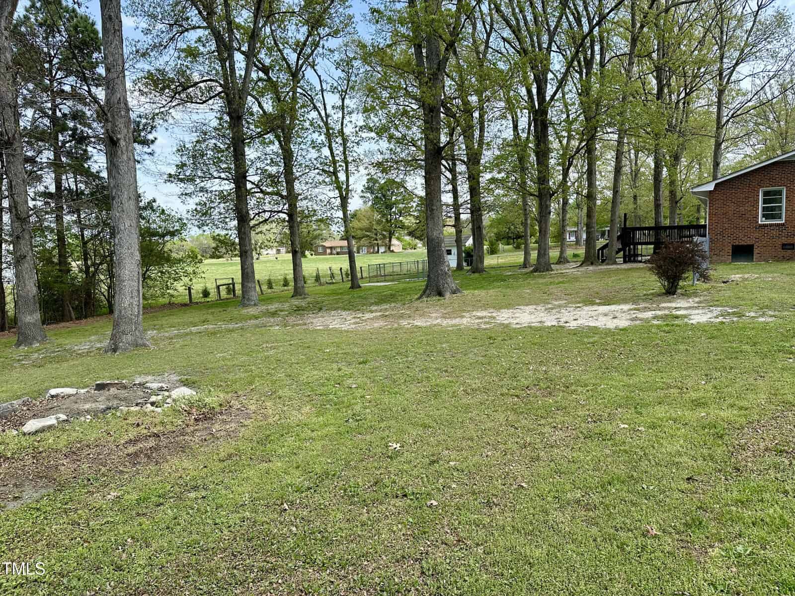 A grassy backyard with scattered trees and a visible dirt patch in the foreground. A brick house is partially visible on the right. In the background, there is a fence, more trees, and two small wooden structures. The sky is cloudy.