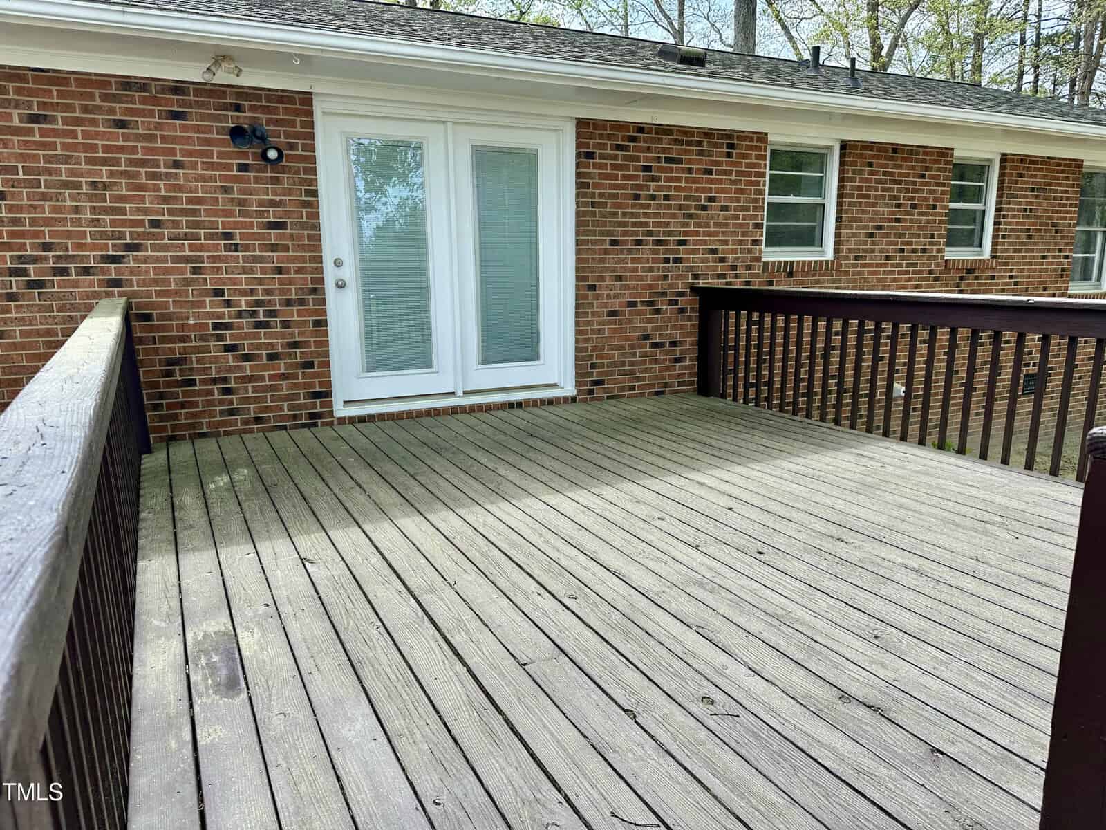 A wooden deck attached to a brick house features a pair of glass double doors. The deck has wooden railings and is situated in front of a two-story brick wall with several windows. The area appears to be outdoors and surrounded by trees.