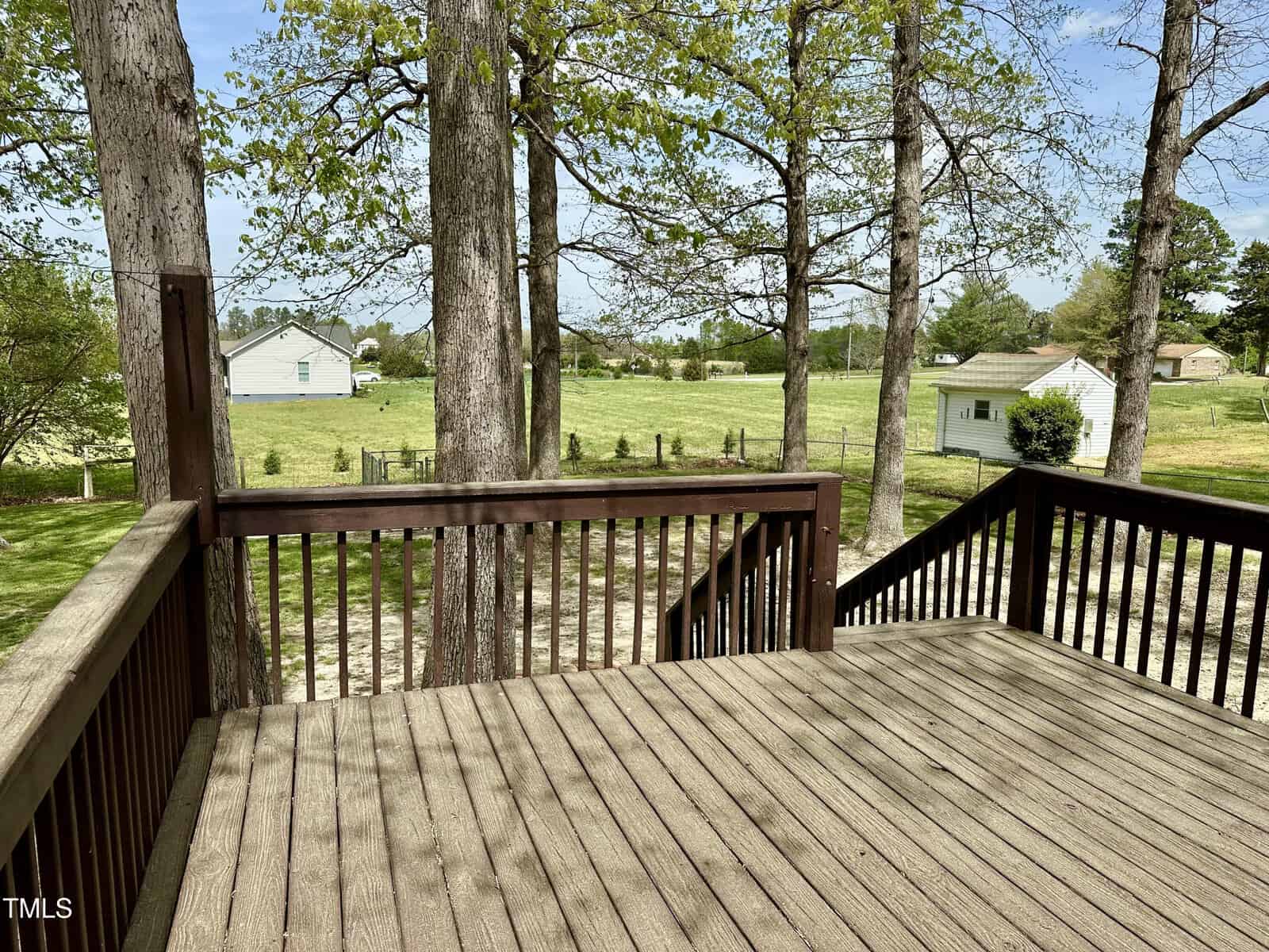 Wooden deck overlooking a grassy yard with several trees. In the background, there are houses surrounded by fields and more trees. The sky is partly cloudy.