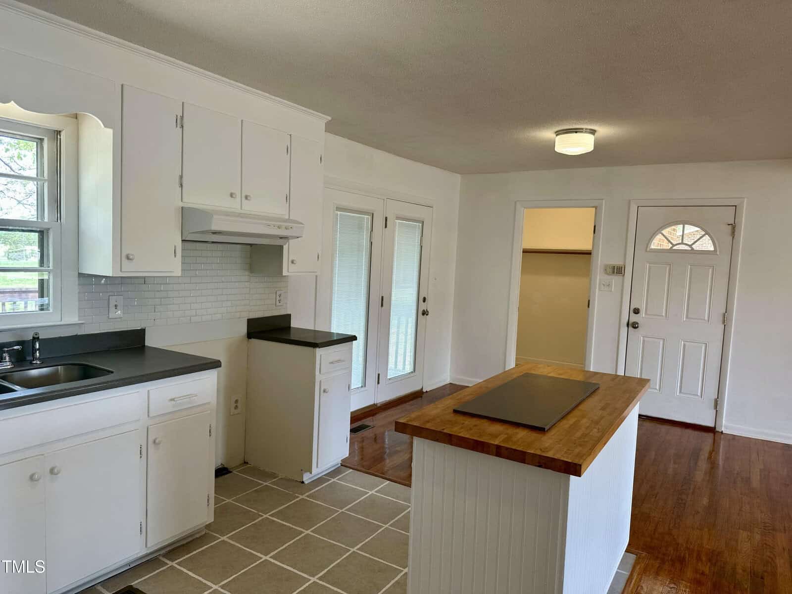 A kitchen with white cabinets, a tiled backsplash, and a dark countertop. An island with a wooden top is in the center. The floor is tiled near the kitchen and wooden near the door. French doors lead outside, and there's a light fixture on the ceiling.