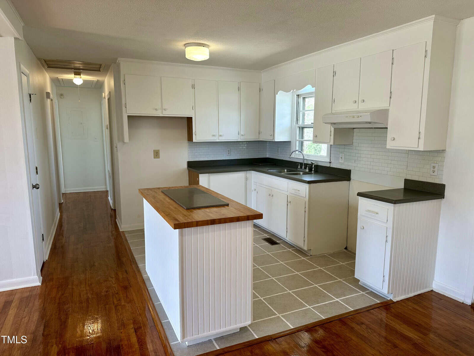 A bright kitchen with white cabinets and a wood-topped island. There is a black countertop, white tile backsplash, stove, and dishwasher. The floor features both wood and tile. A hallway extends from the kitchen.