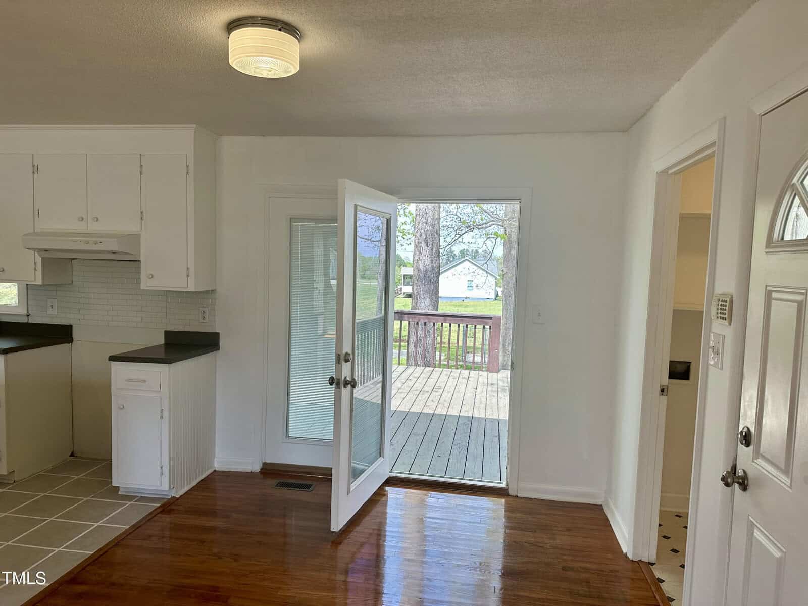 A room with white walls features a partial kitchen view, including white cabinets and hood. A door with glass panels opens to a wooden deck. There is a view of the outside lawn and a neighboring house. Wood and tile flooring are visible.