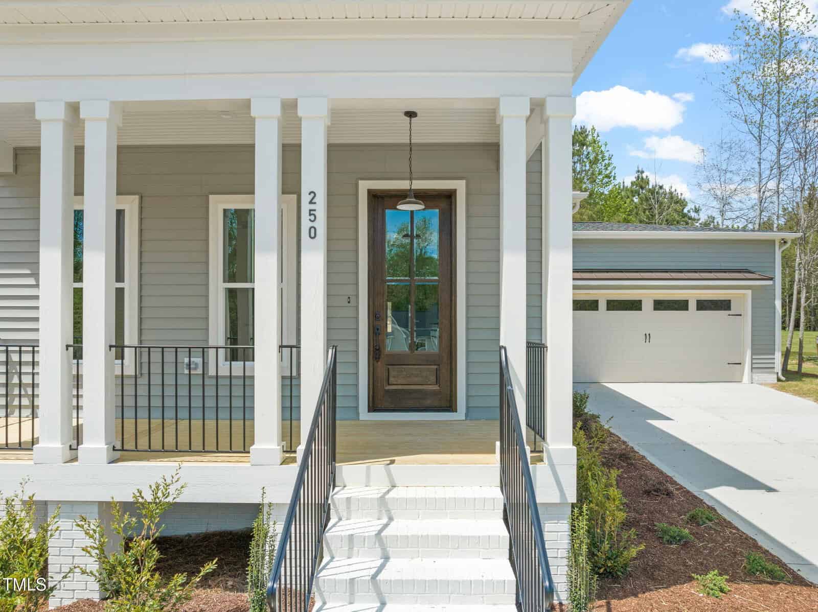Front view of a house showing a covered porch with white columns, a wooden front door with glass panel, black metal railing, and steps leading to the entrance. A detached two-car garage is visible in the background.