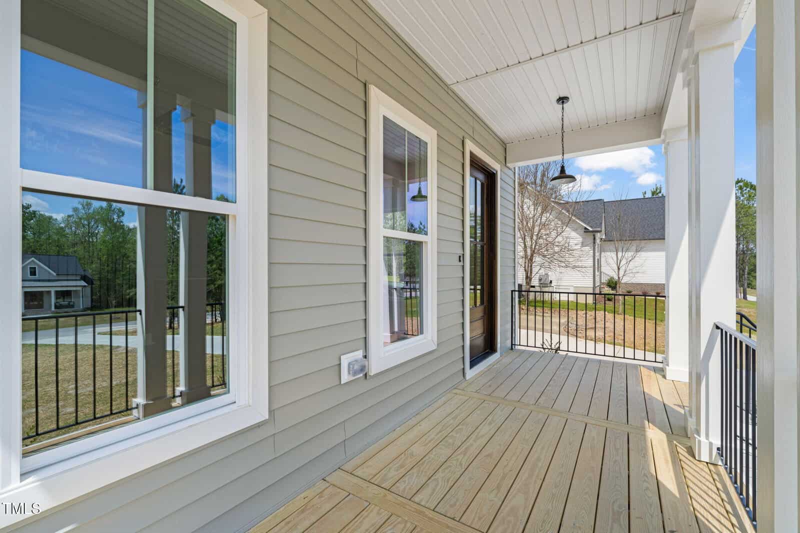 A wooden front porch with beige siding, white trim, black metal railing, and two windows. There is a ceiling light fixture, a brown door, and a view of a suburban street and neighboring houses.