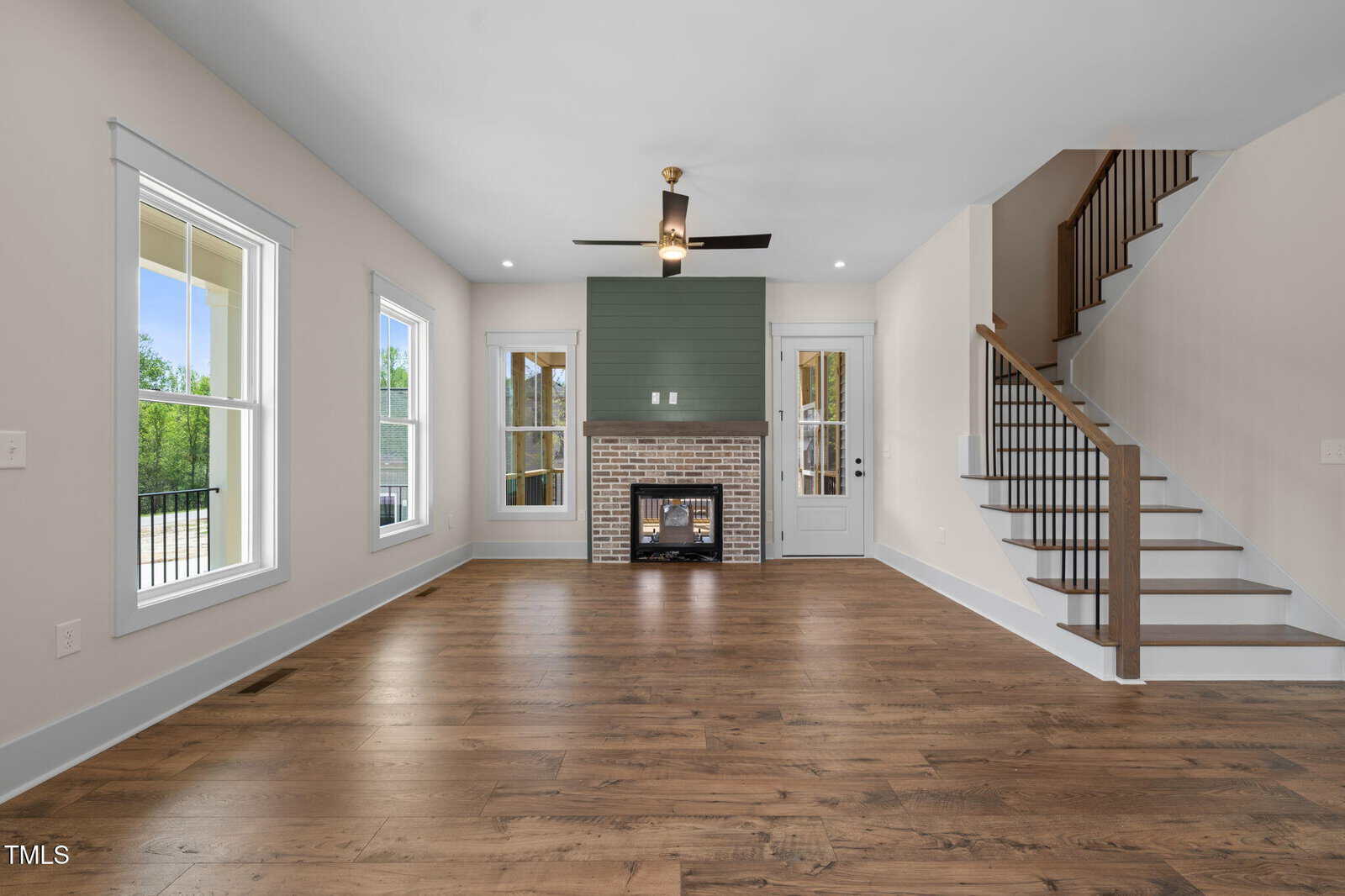 A modern living room with wood flooring, white walls, large windows, a ceiling fan, a brick fireplace with a green accent wall, and a staircase with a wooden handrail leading upstairs.