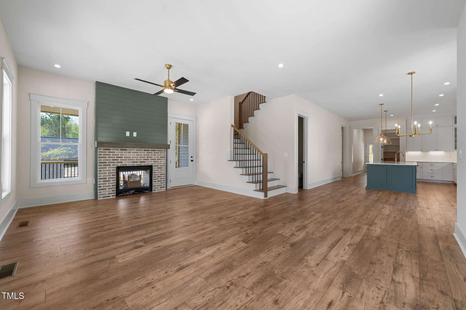 Spacious open-concept living area with wood floors, a brick fireplace, ceiling fan, staircase, large windows, and a view into the kitchen with white cabinets and a blue island.