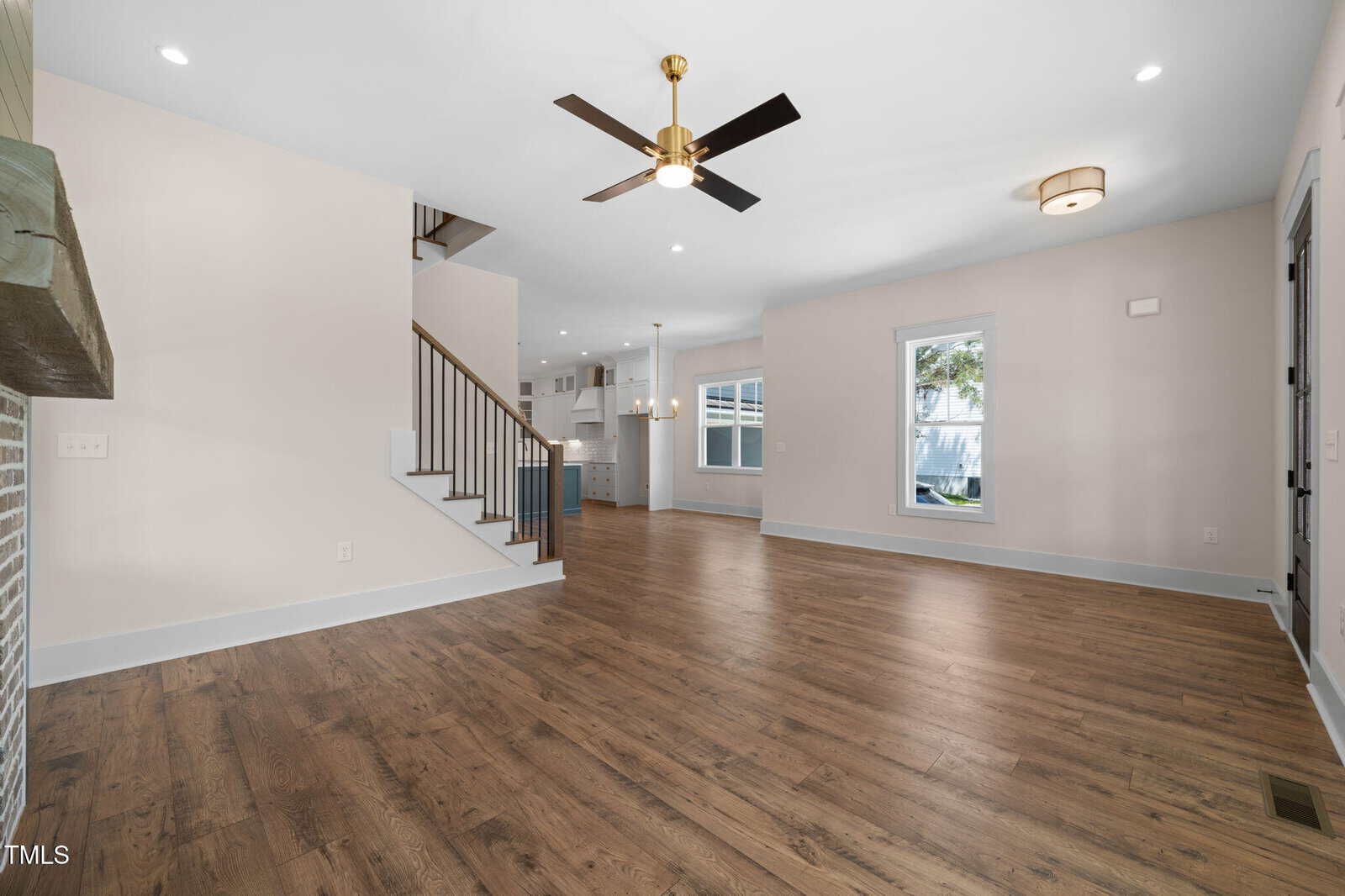 Spacious living area with hardwood floors, white walls, a ceiling fan, recessed lighting, and visible staircase; kitchen with white cabinets and pendant lights is seen in the background. Large windows provide natural light.