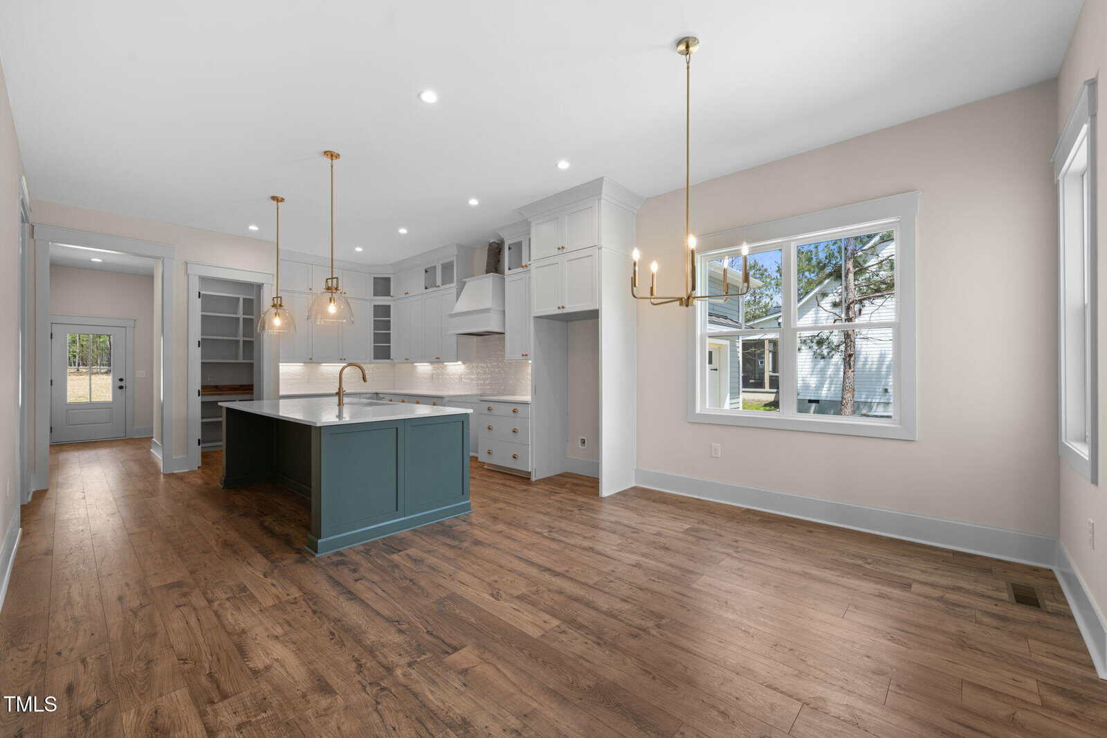 Modern kitchen with wood flooring, a blue island with a sink, white cabinets, pendant lights, built-in shelving, and large windows letting in natural light. Adjoining room visible through an open doorway.