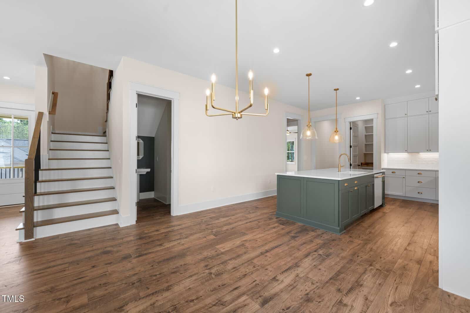 Modern kitchen with wood floors, a green island with a white countertop, gold light fixtures, white cabinetry, and a staircase to the left. A small bathroom is visible under the stairs. The space is empty and well-lit.