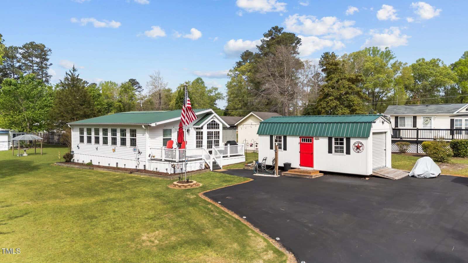 A white single-story house with a front deck and an American flag stands next to a green-roofed shed with a red door. There is a paved driveway and a grassy yard surrounded by trees under a partly cloudy sky.