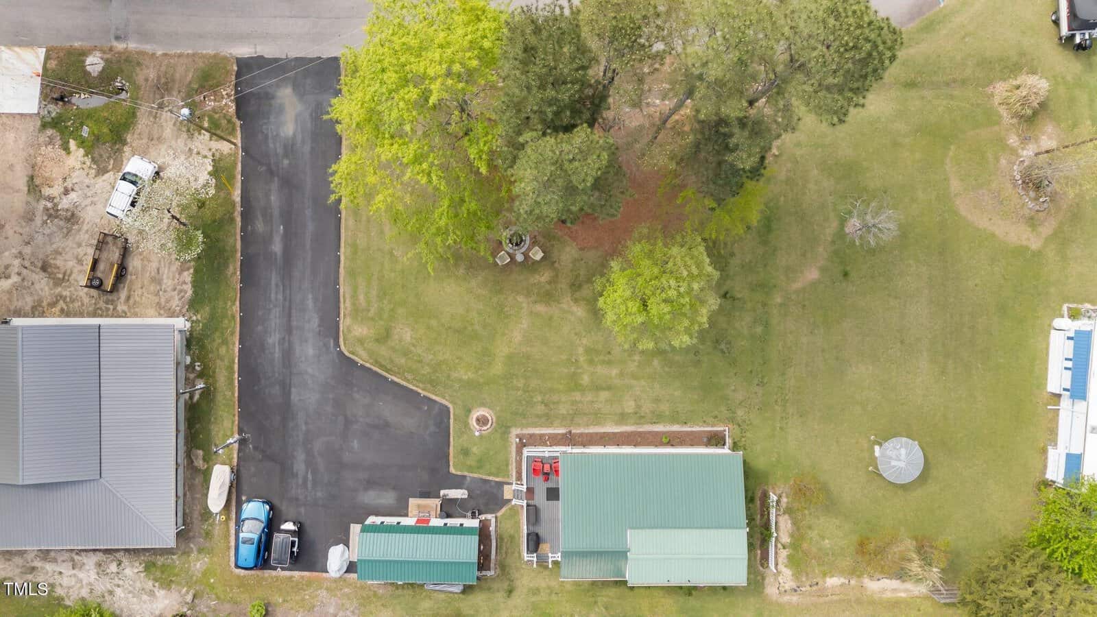 Aerial view of a property with two green-roofed buildings, paved driveway, parked vehicles, and grassy areas with trees. Adjacent structures and streets are partially visible at the edges of the image.