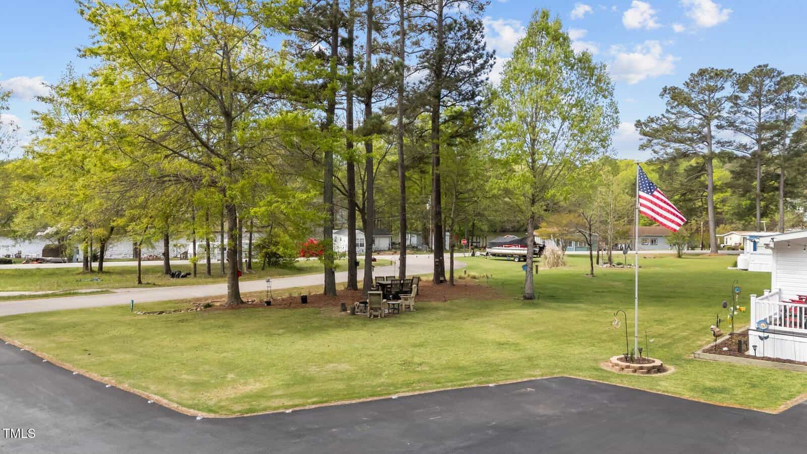 A grassy yard with tall trees, an American flag on a pole, outdoor seating around a fire pit, and a paved driveway. Houses and a lake are visible in the background under a blue sky with scattered clouds.