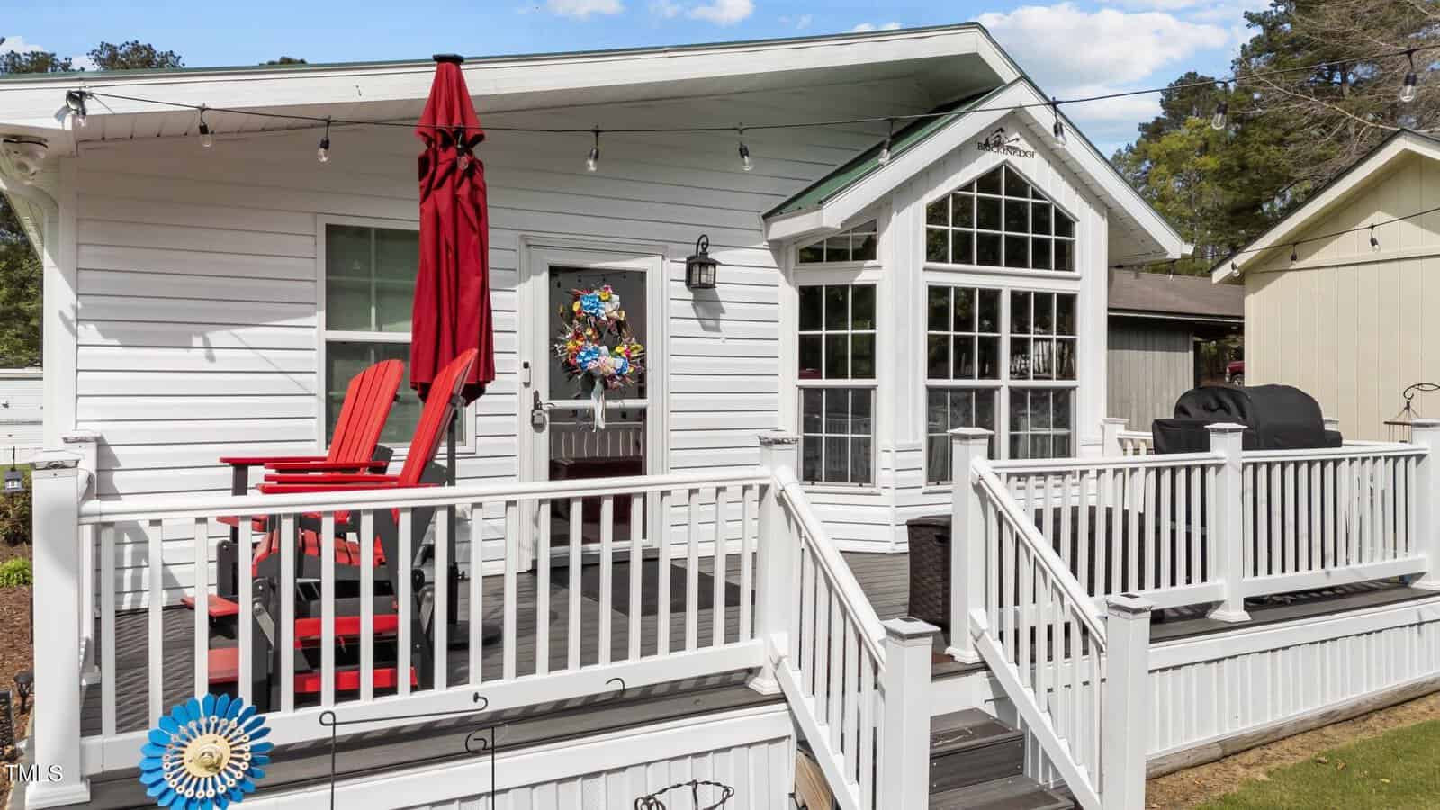 A small white house with a front porch featuring red Adirondack chairs, a red patio umbrella, a grill, and a wreath on the door. String lights hang above, and a blue ribbon is attached to the porch railing.