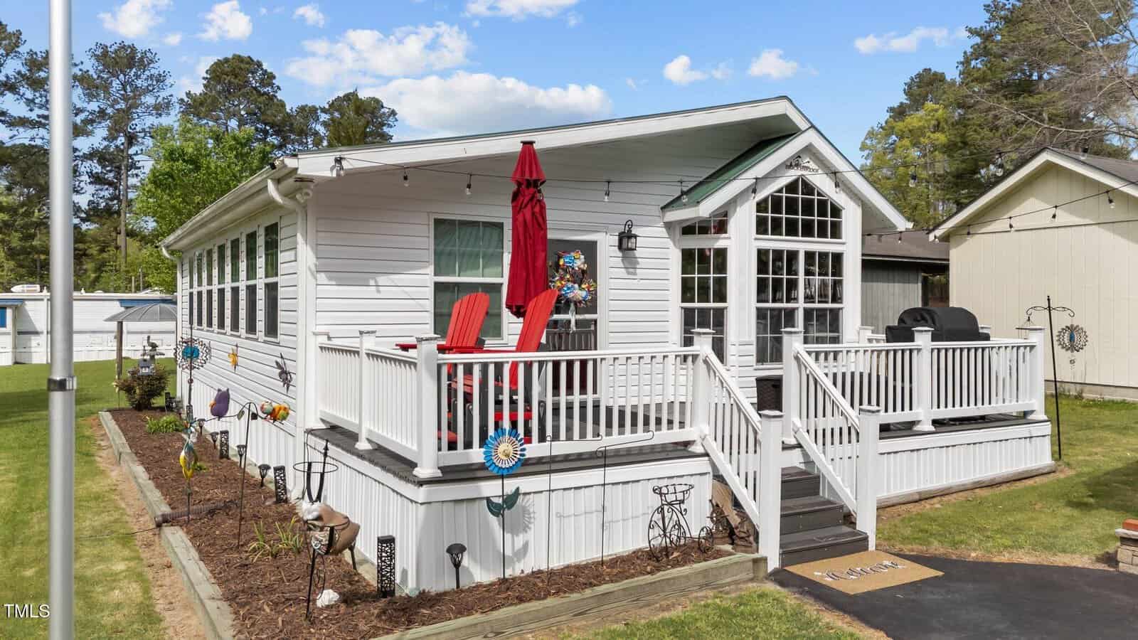 A white single-story house with a raised porch featuring red chairs, a red umbrella, and a grill. The porch is surrounded by a white railing and overlooks a landscaped yard with flowers and garden decorations.