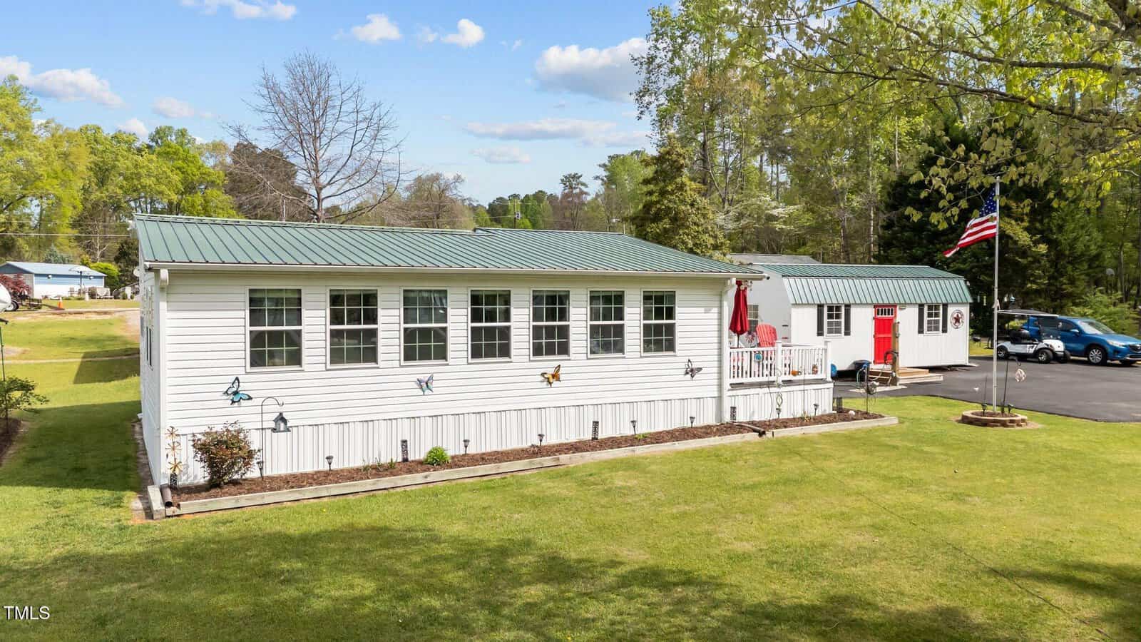 A white single-story house with a green metal roof, butterfly decorations on the exterior, a front porch with red accents, an American flag, and a well-kept lawn with trees and a paved driveway.