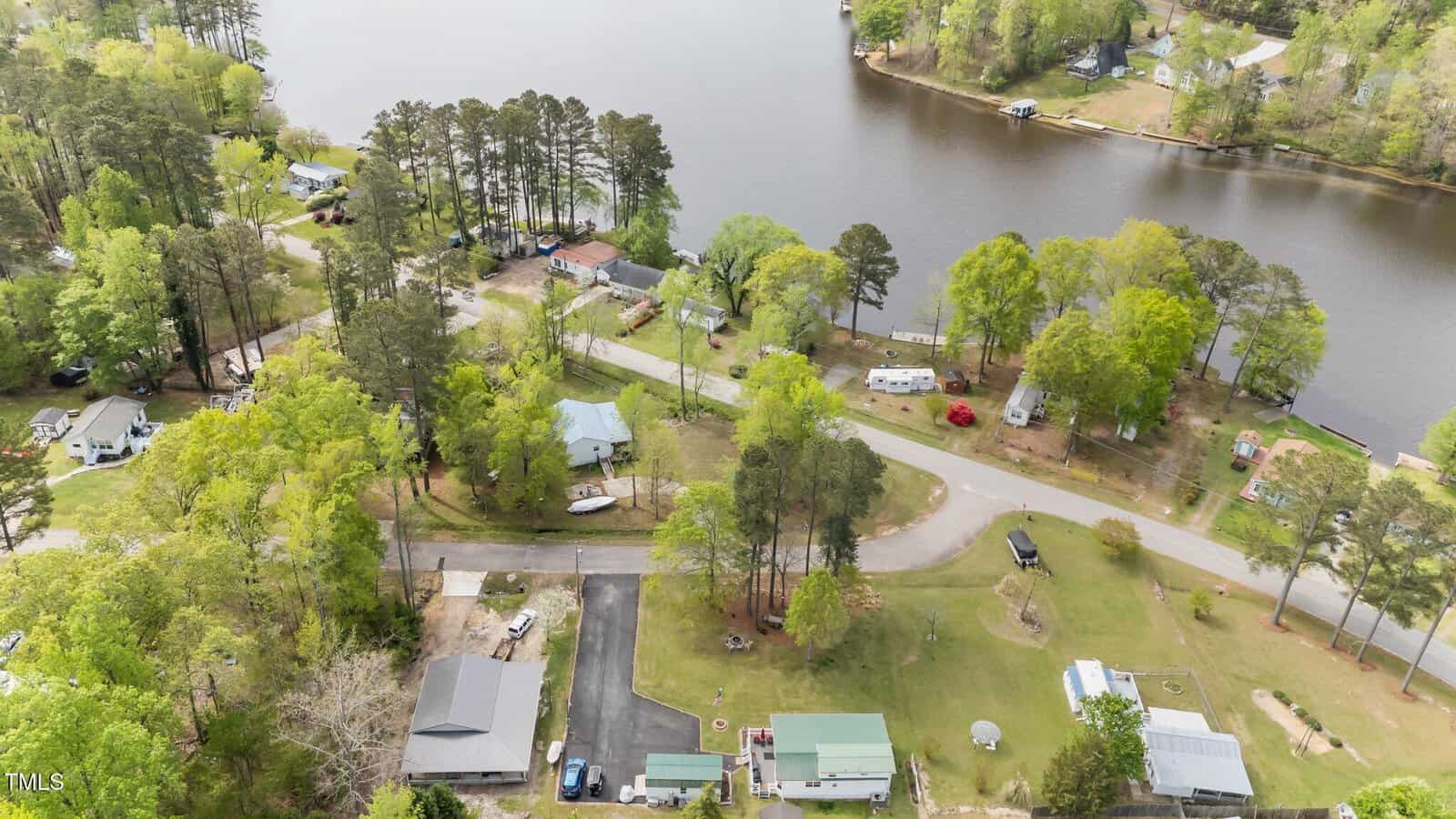 Aerial view of a small lakeside community with houses, roads, scattered vehicles, trees, and grassy areas near a body of water, with a curved road running along the shoreline.