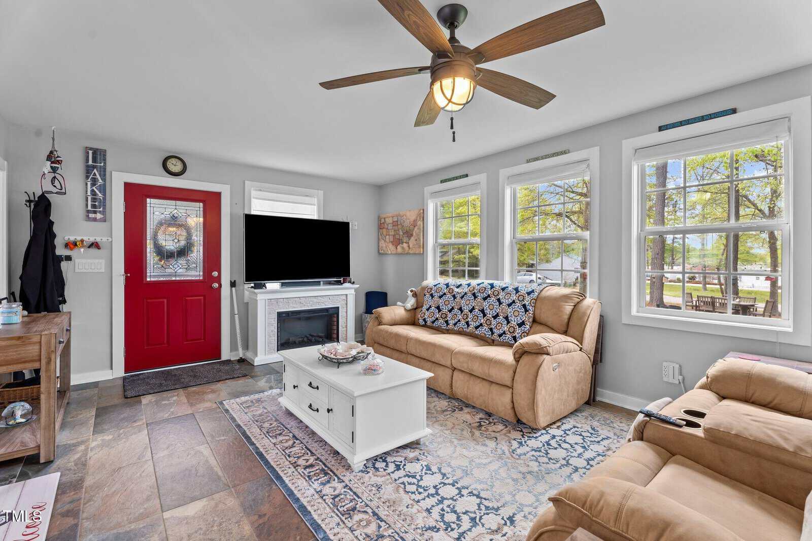 A living room with a red door, tan sofas, a ceiling fan, large windows, a TV above a fireplace, a white coffee table, a patterned rug, and tile flooring. Decor includes wall art and a coat rack near the door.