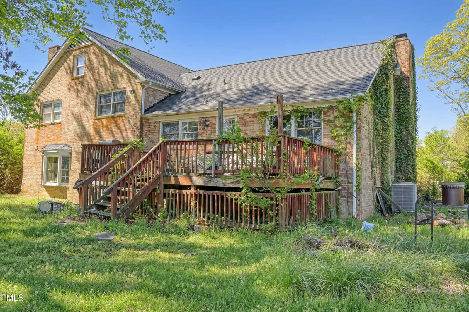 A brick house with ivy growing on one side features a wooden deck at the back. The deck has a railing and stairs leading down to a grassy yard. The sky is clear, and trees surround the property, creating a natural setting.