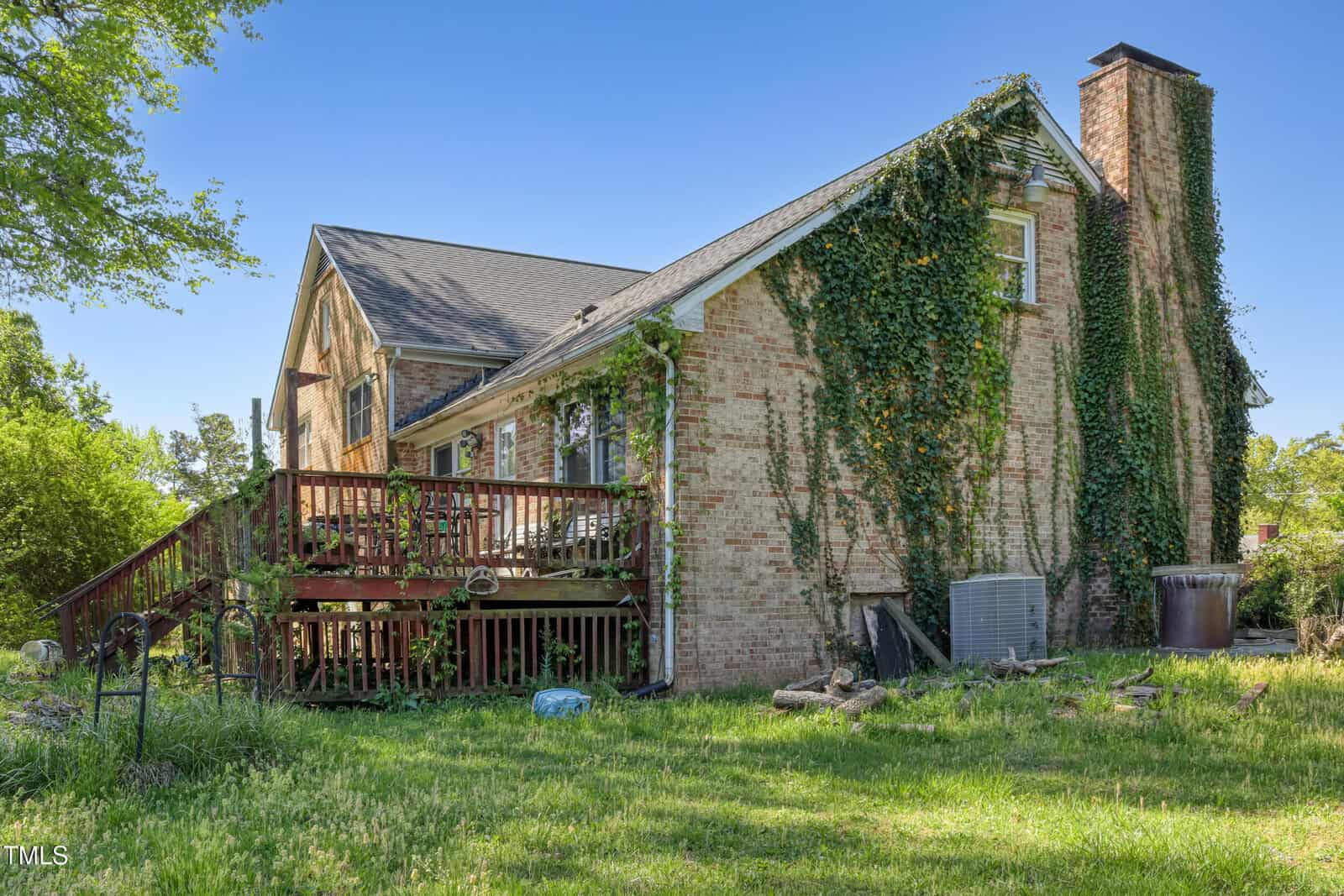A brick house with a steep roof is partially covered in ivy. A wooden deck extends from one side of the house. The yard is grassy, and there is a visible air conditioning unit and some scattered objects on the lawn.