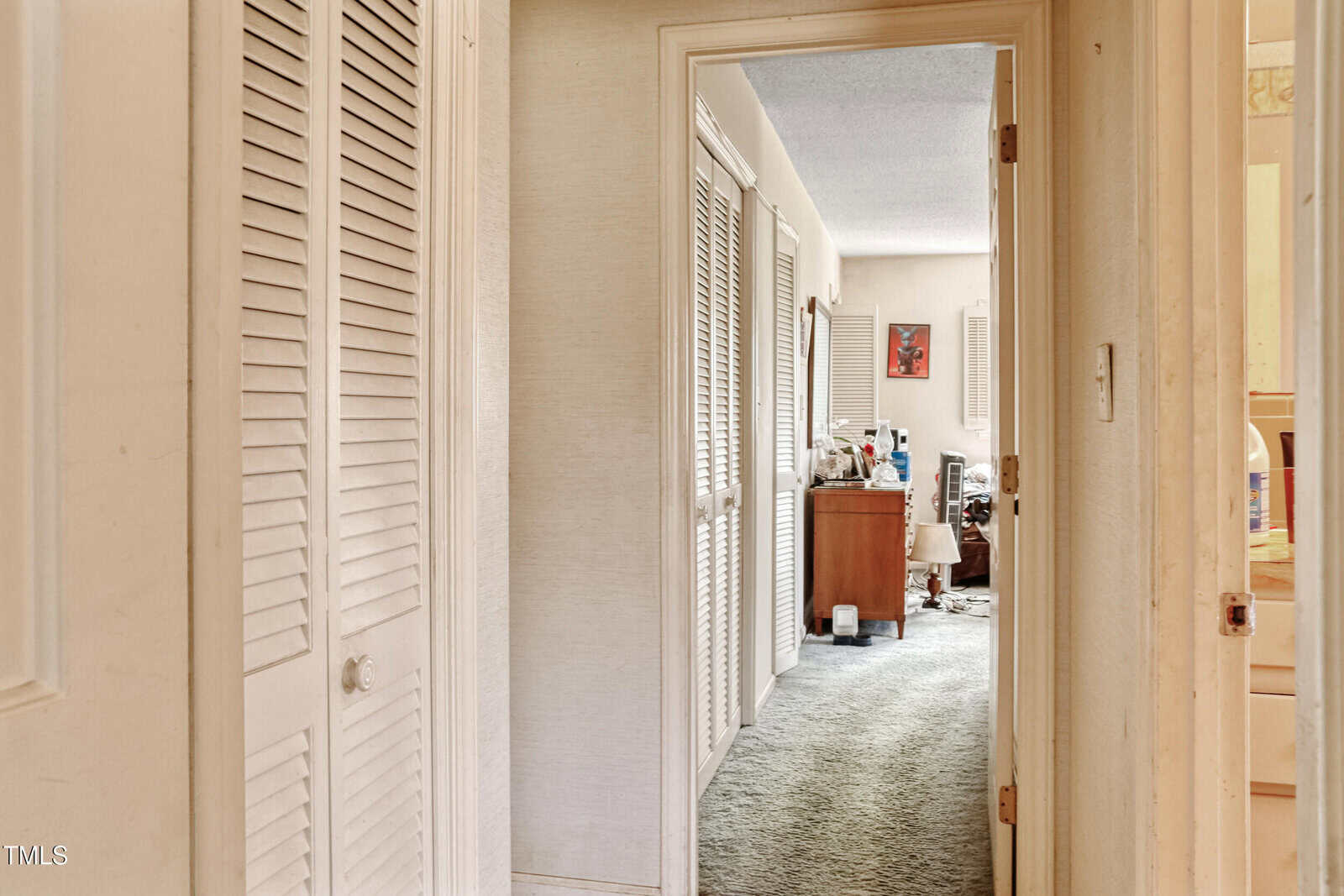 A hallway with white louvered doors on the left leads to a room with green carpet. The room contains a wooden desk, a chair, a cluttered tabletop, and a bright red picture on the wall. The right has a partially visible doorway.